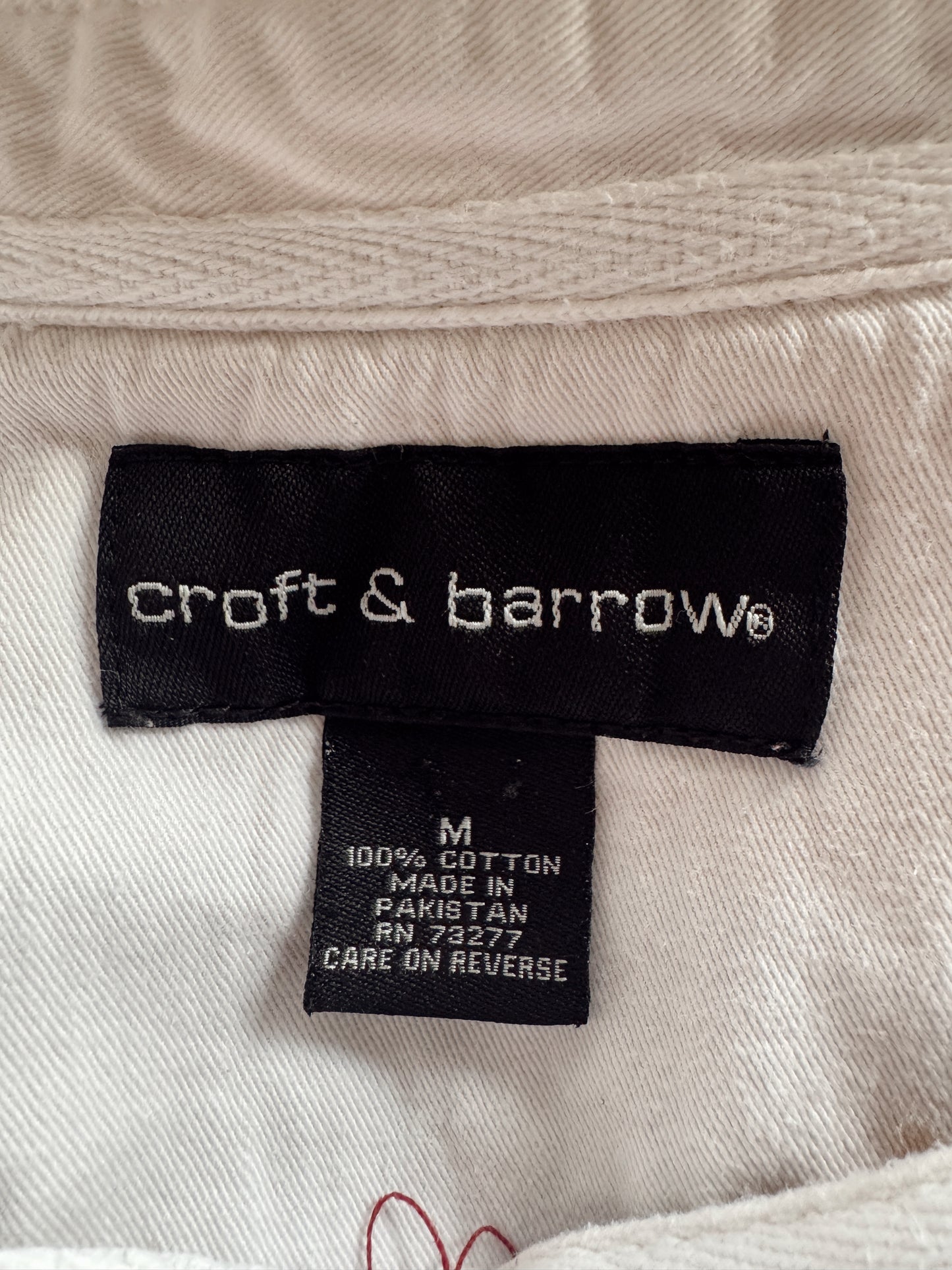 Close-up of a clothing label reading 'croft & barrow' on a light beige fabric garment. A smaller tag below shows size M, 100% cotton, made in Pakistan.