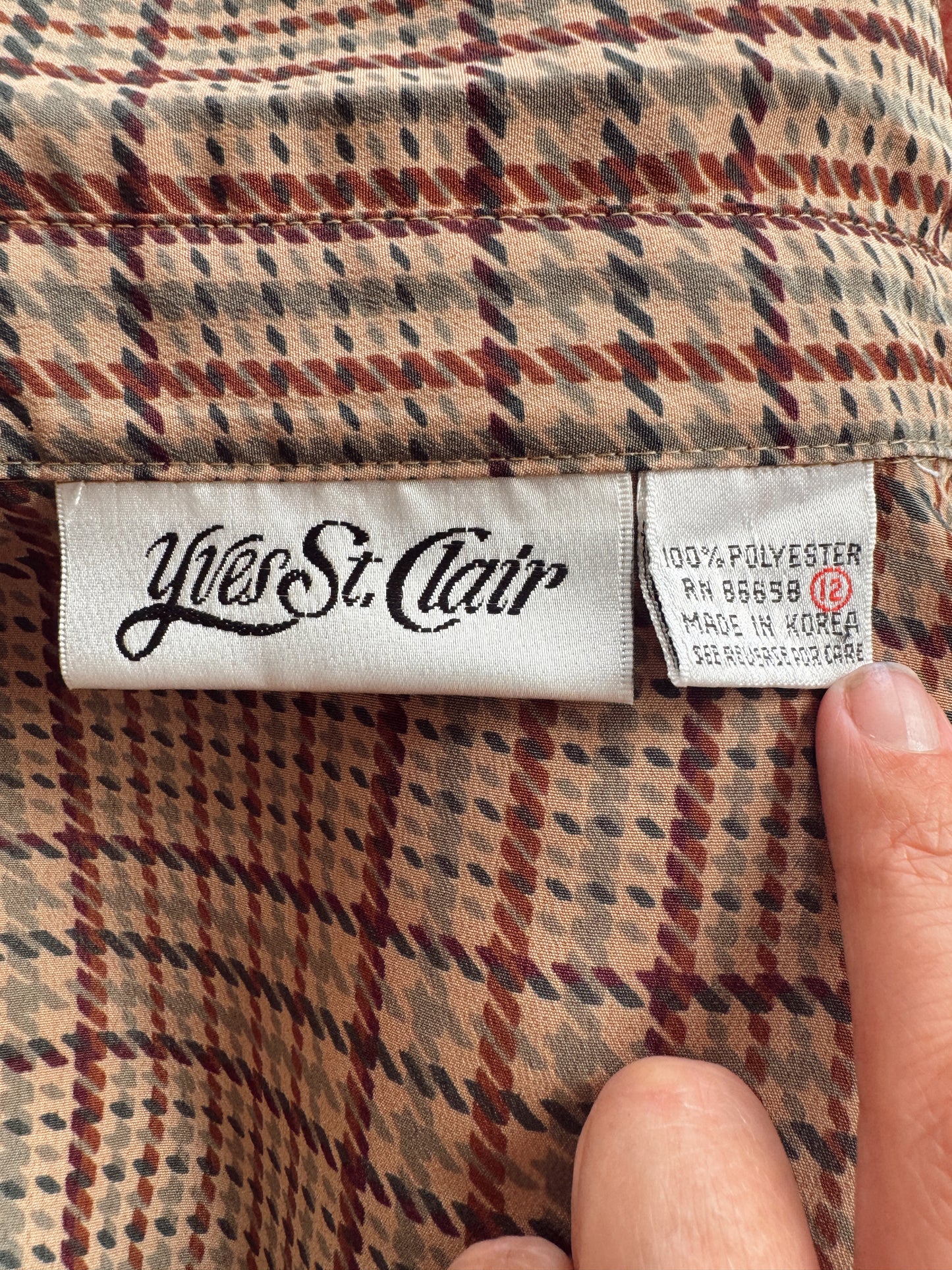 Close-up of a plaid garment label reading Yves Saint Clair, with a finger pointing at the fabric. The visible textile is beige, brown, and red plaid.