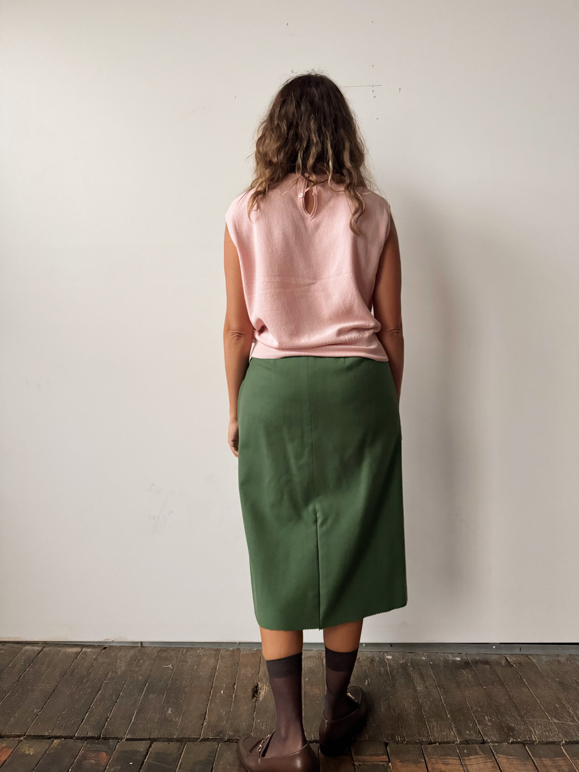 Person wearing a pink sleeveless top and green skirt against a white wall.