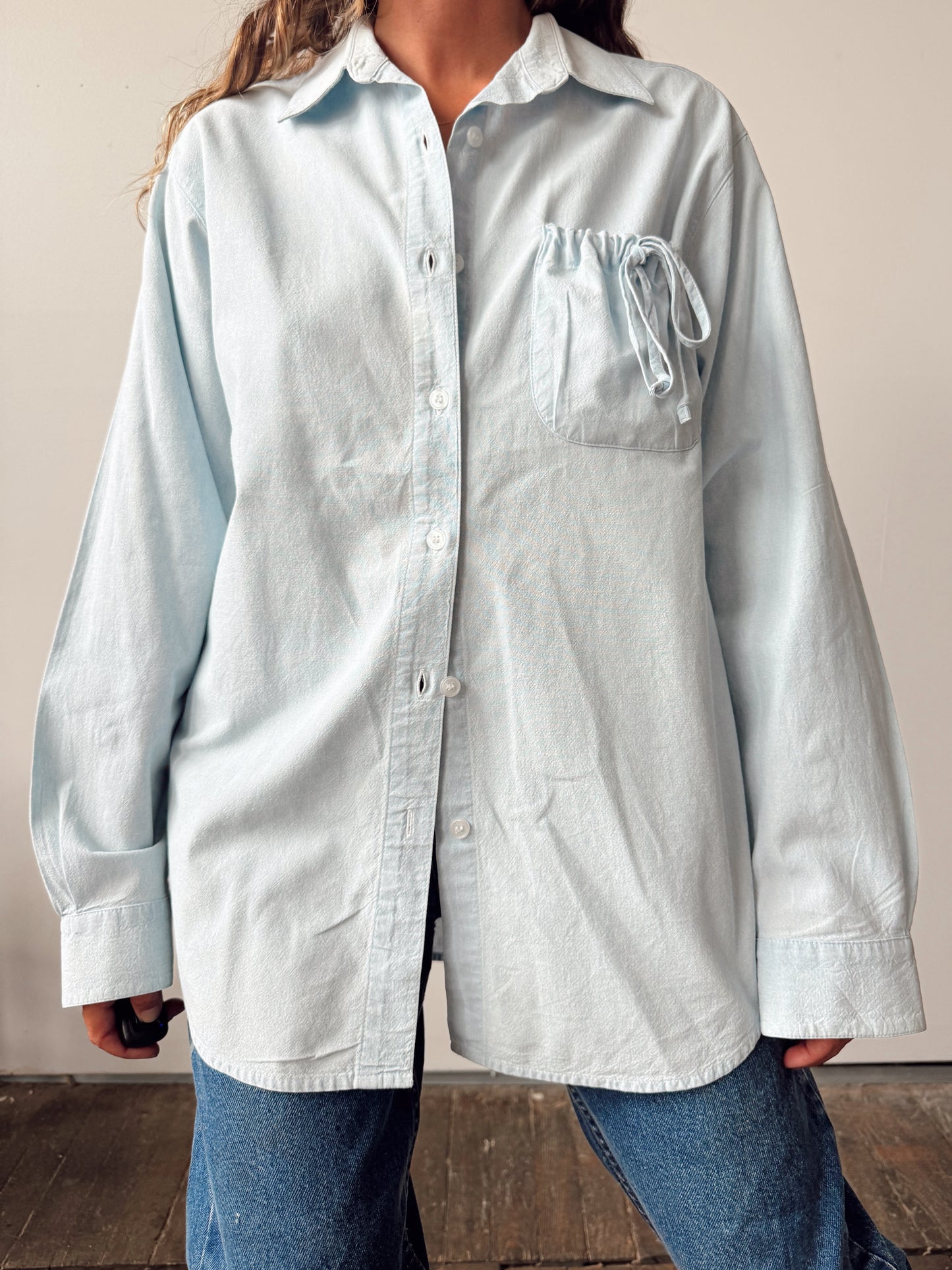 A person wearing an oversized light blue button-up shirt with a chest pocket and drawstring detail, paired with blue jeans, standing against a plain indoor wall.