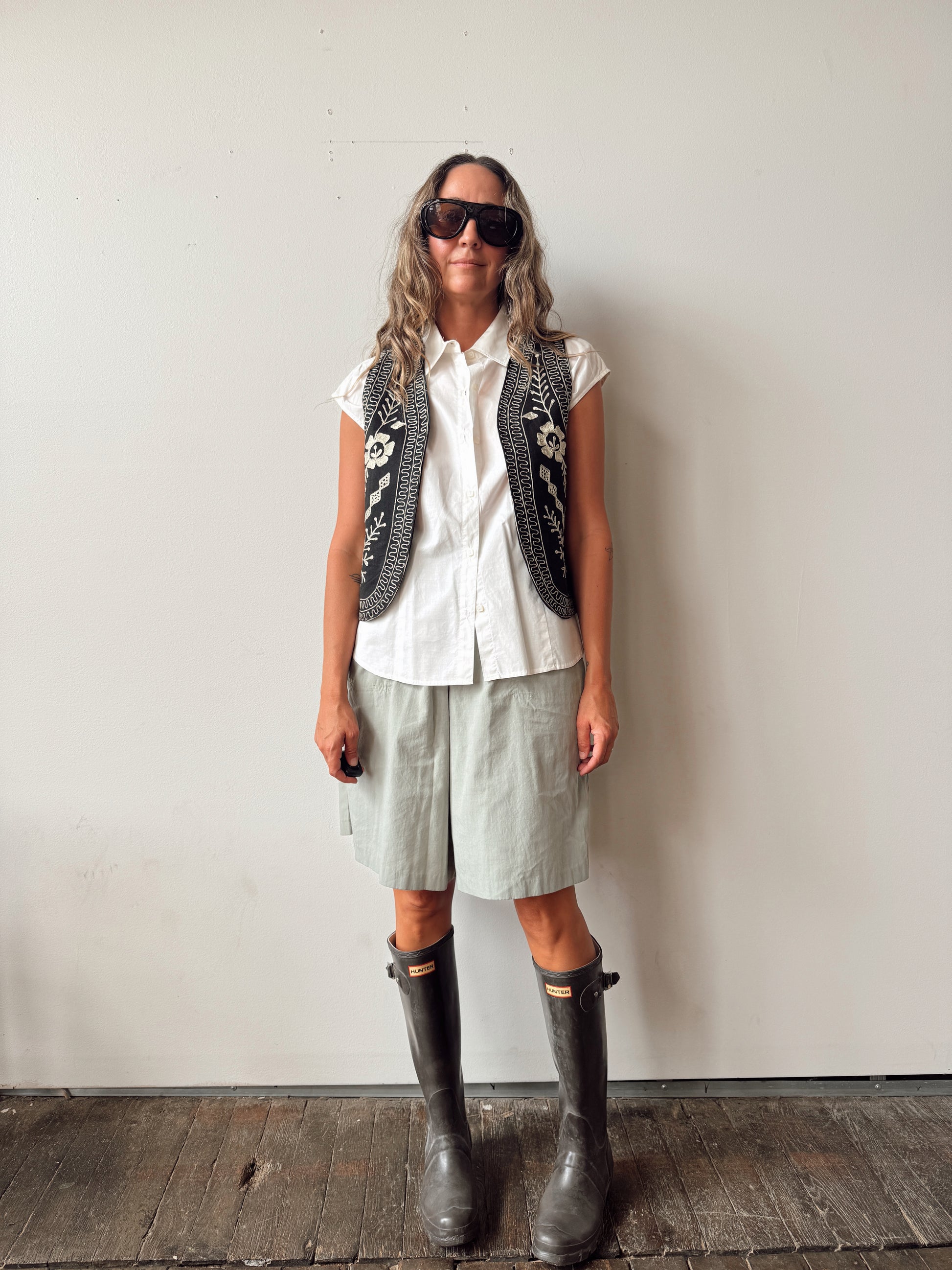 A woman stands against a plain off-white wall wearing a white short-sleeve button-up shirt, a black embroidered vest, light sage green shorts, and dark gray rain boots.