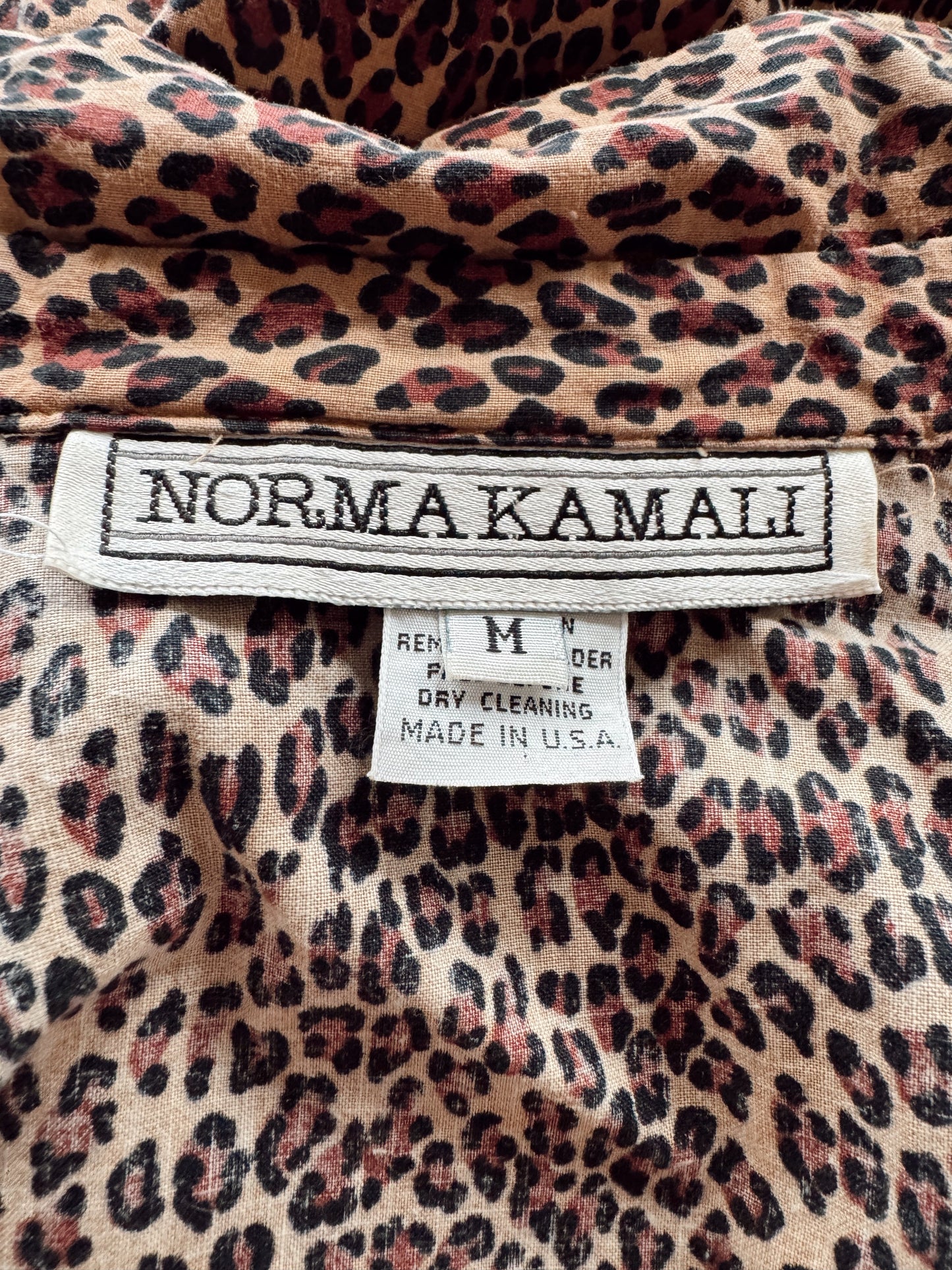 Close-up of a leopard print garment interior showing a size and care label. The fabric has a beige base with black and dusty rose-brown leopard spots.