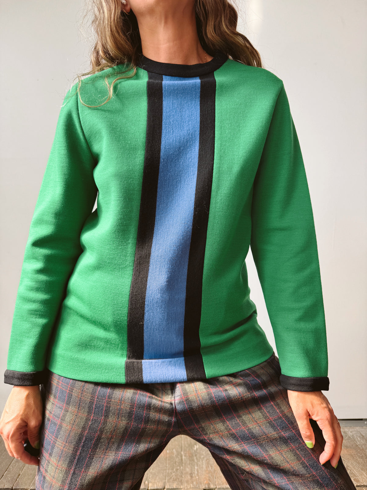 A woman wearing a green knit sweater with a vertical color-blocked design featuring black stripes and a central light blue stripe, paired with plaid trousers.