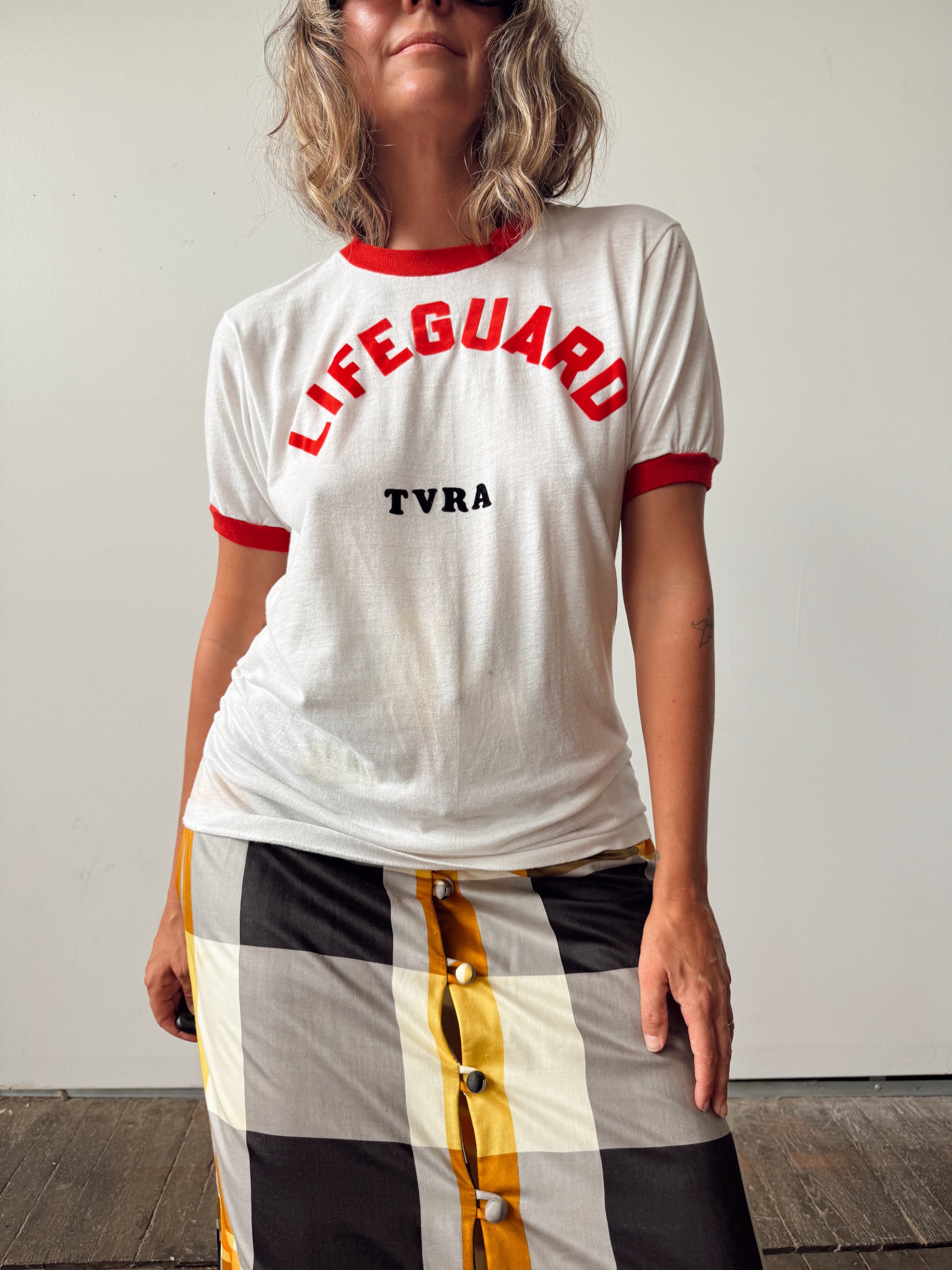 A woman wearing a white ringer T-shirt with red trim and 'LIFEGUARD' printed in red, plus 'TVRA' in black. She is also wearing a black, white, and yellow checkered skirt with gold buttons.