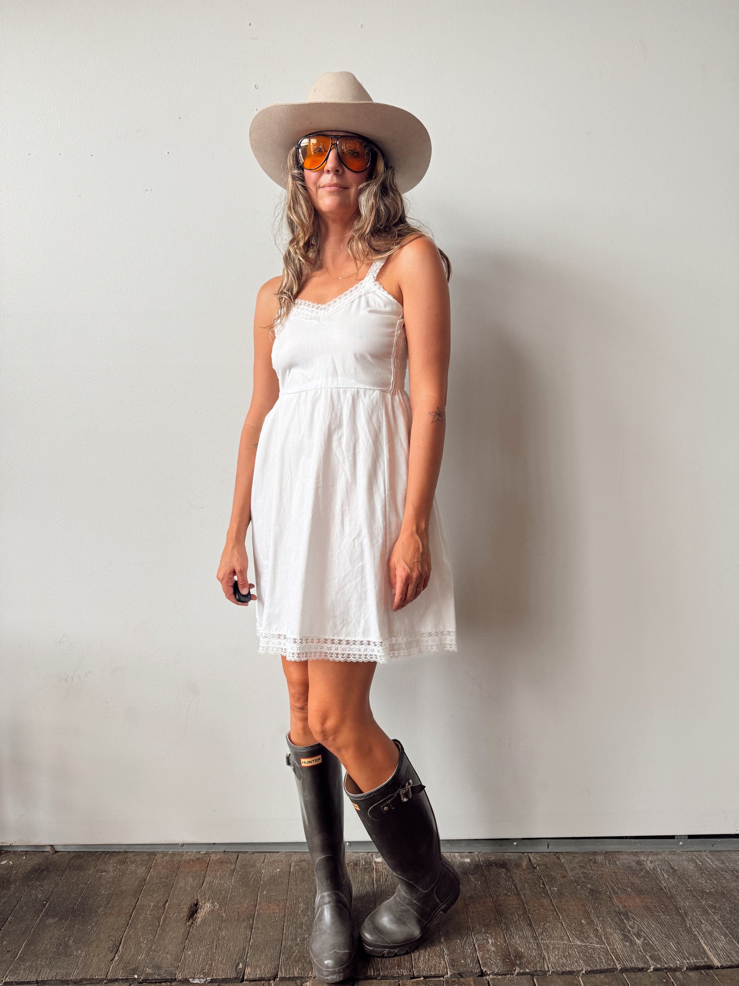 A woman wearing a white sleeveless mini dress, a beige cowboy hat, orange-tinted sunglasses, and black rain boots, standing against a plain light gray wall on a dark wooden floor.