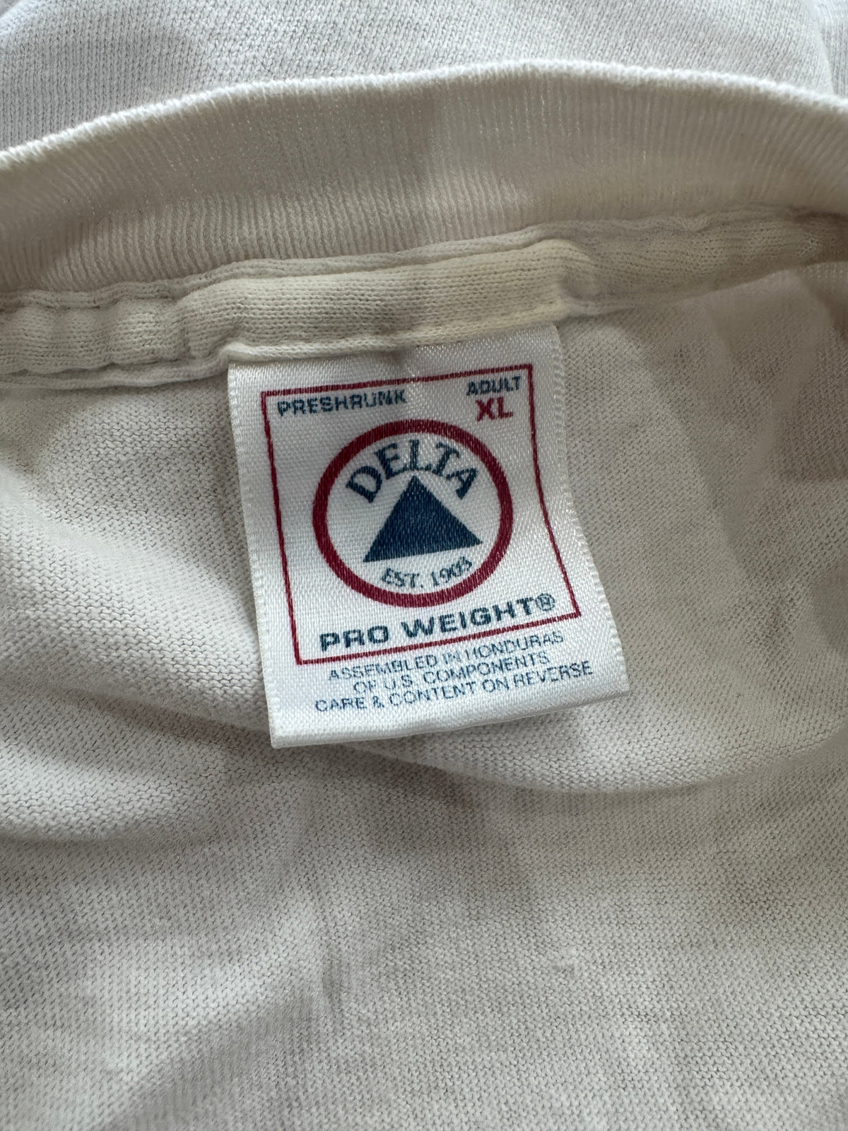 Close-up of a white fabric garment tag with the Delta brand logo and 'Pro Weight' text, sewn onto an off-white ribbed knit garment.