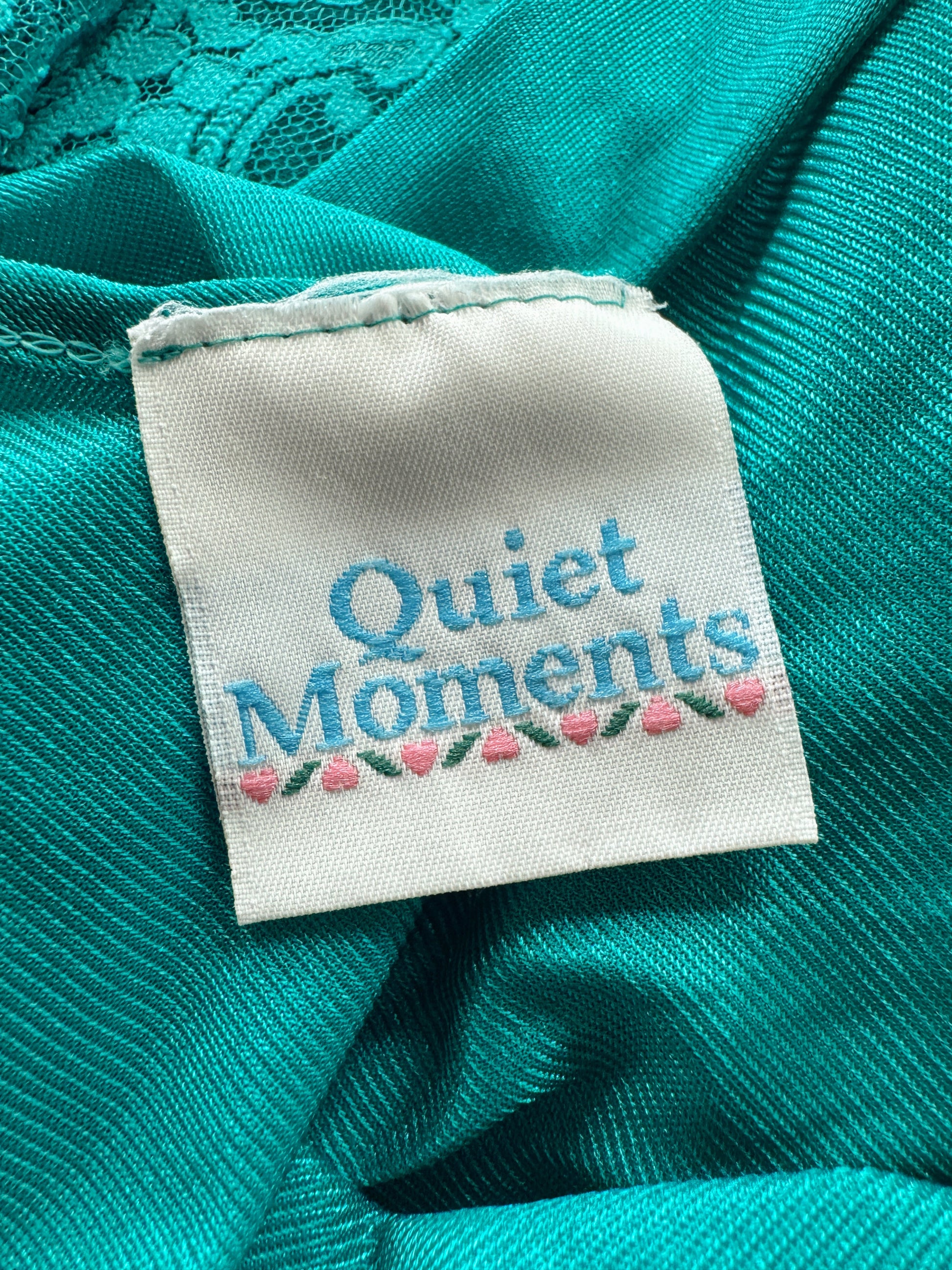 A close-up of a white fabric sewn-in label reading 'Quiet Moments' in blue lettering with pink flowers and green leaves, attached to teal-green textured fabric.
