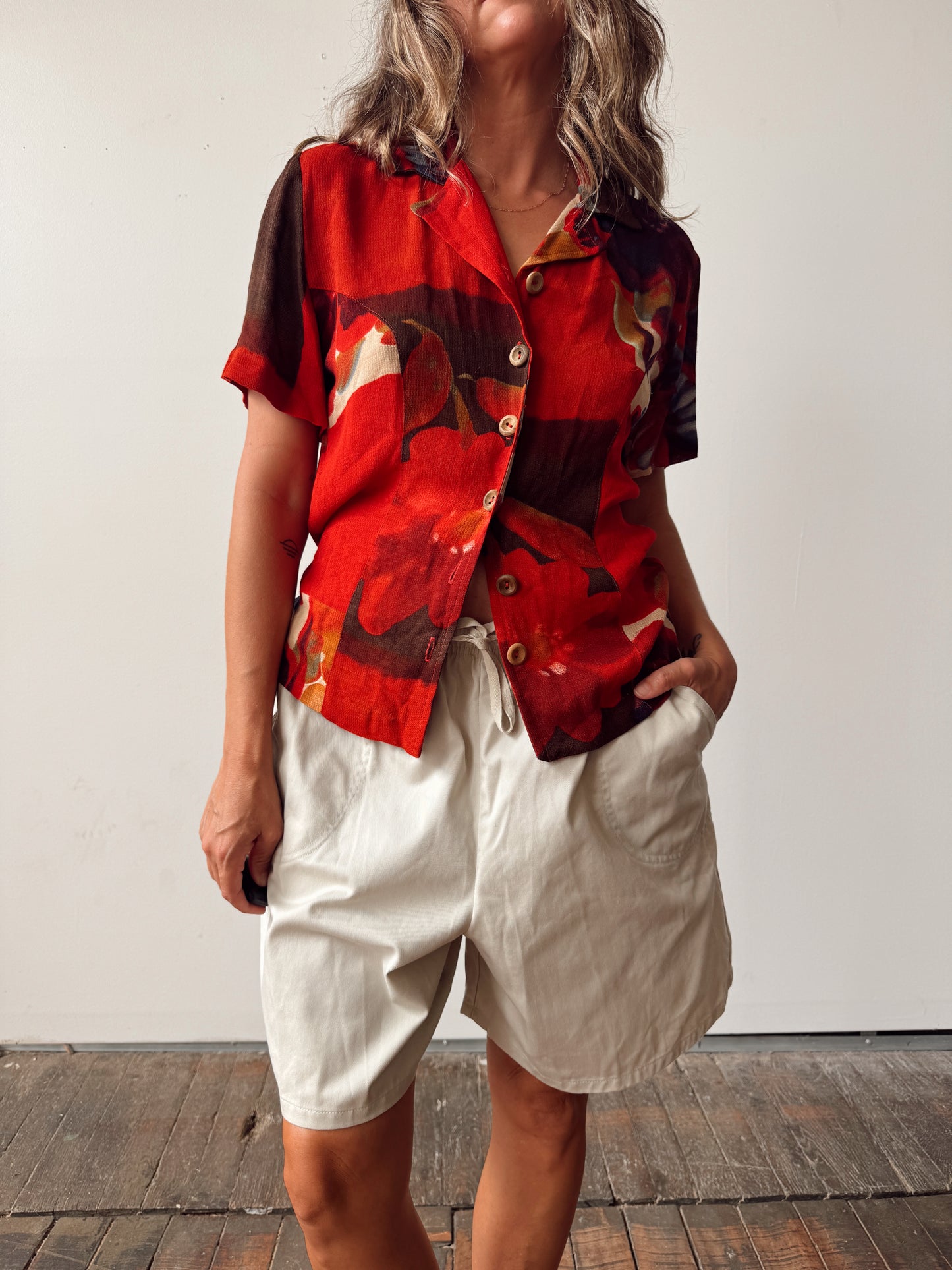 A woman wearing a red and black abstract print short-sleeve button-up shirt with cream shorts, standing against a plain off-white wall.