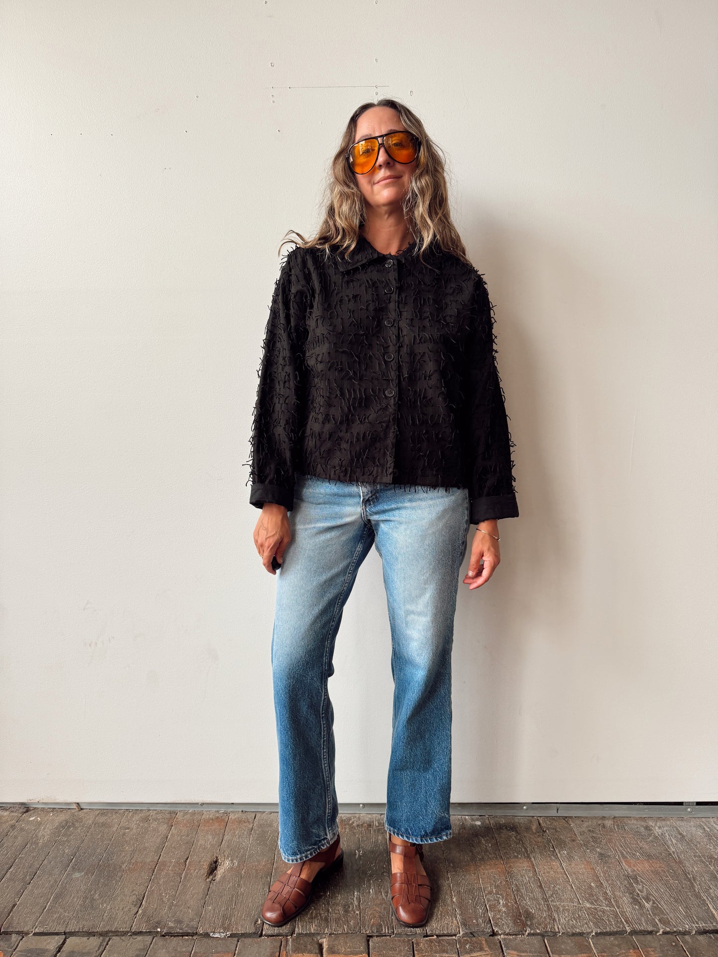 A woman stands against a plain off-white wall wearing a black fuzzy cropped jacket, light blue jeans, and brown leather boots. She is also wearing orange-tinted sunglasses.