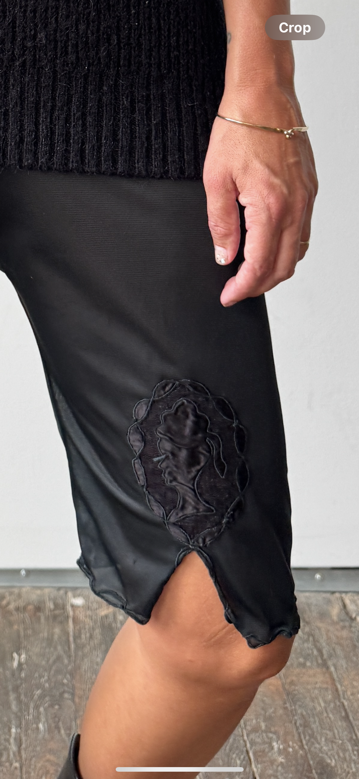 A close-up of a person wearing a black satin-like skirt with a large black embroidered applique patch near the hem and a side slit, paired with a black ribbed top. A gold bracelet is visible on the wrist.