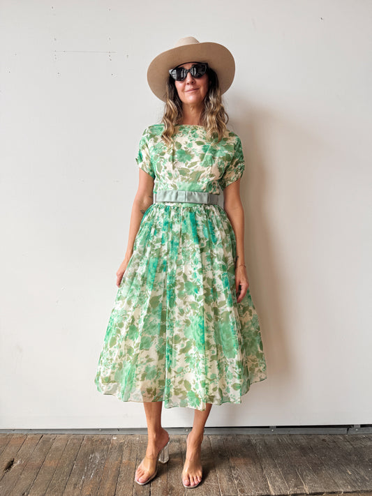 Woman wearing a green floral dress with a hat and sunglasses against a white wall.
