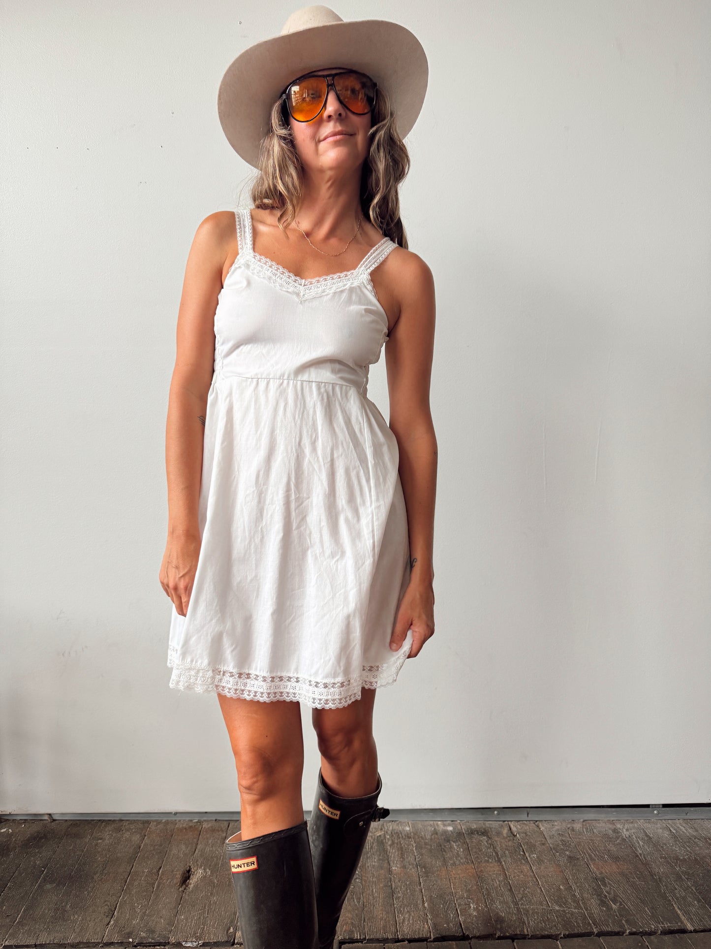 A woman wearing a white sleeveless lace-trimmed dress, a wide-brimmed beige hat, orange-tinted sunglasses, and black rain boots, standing against a plain white wall.