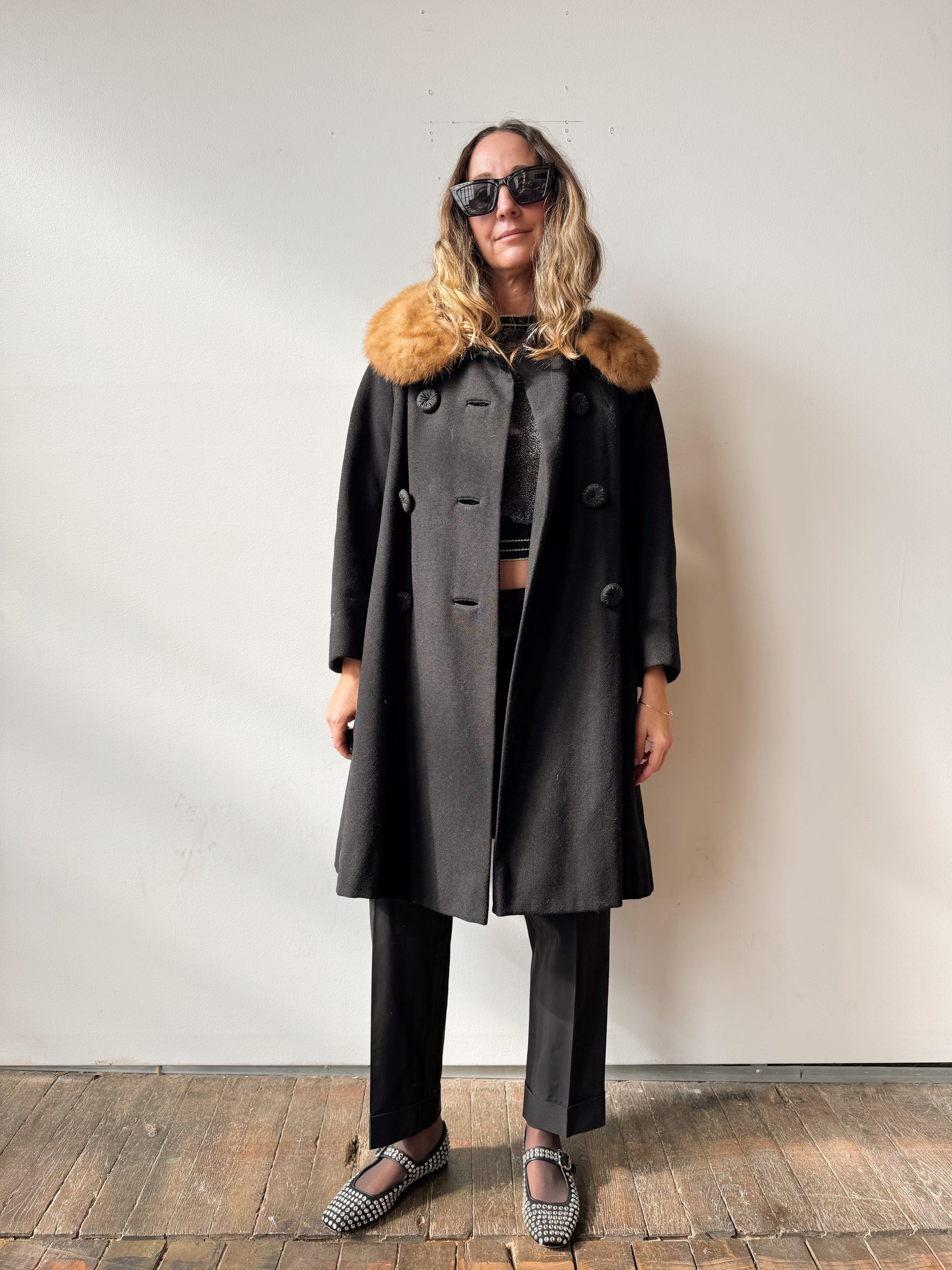 Person wearing a long dark coat with a fur collar against a plain background