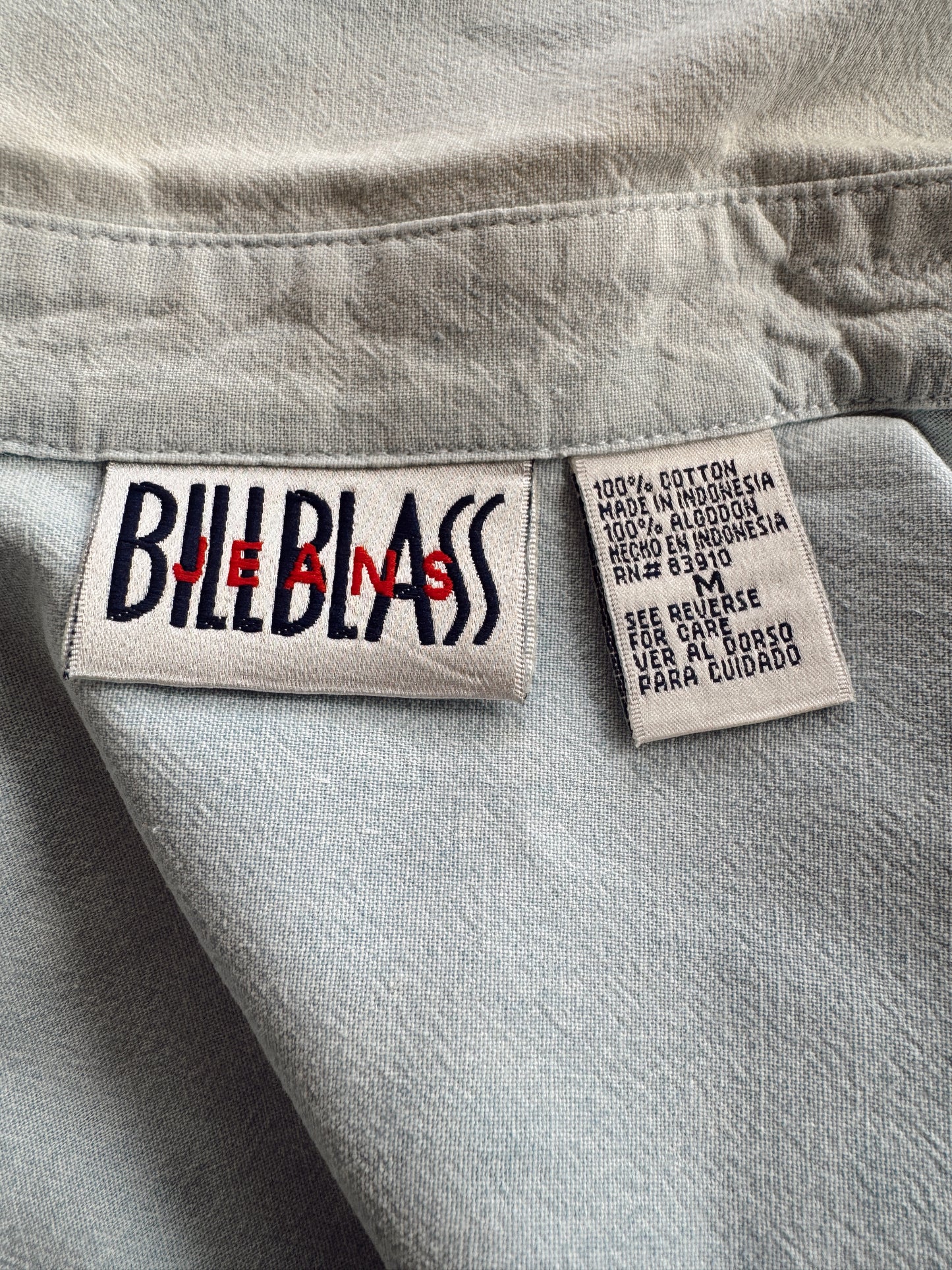 Close-up of a light gray denim garment showing two sewn-in labels: a Bill Blair Jeans logo patch on the left and a material/care label on the right.