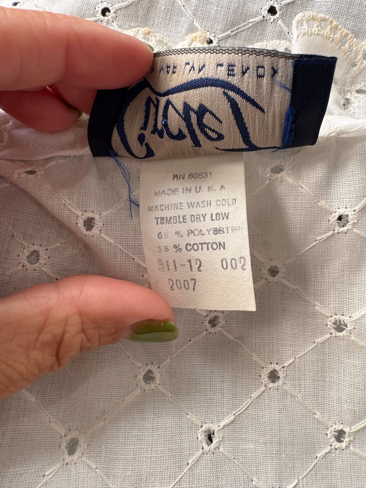 A close-up of a hand holding the inside neck label of a white embroidered blouse. The brand name 'Cactus' is clearly visible on the dark blue fabric label, and the white paper tag shows details including '3.