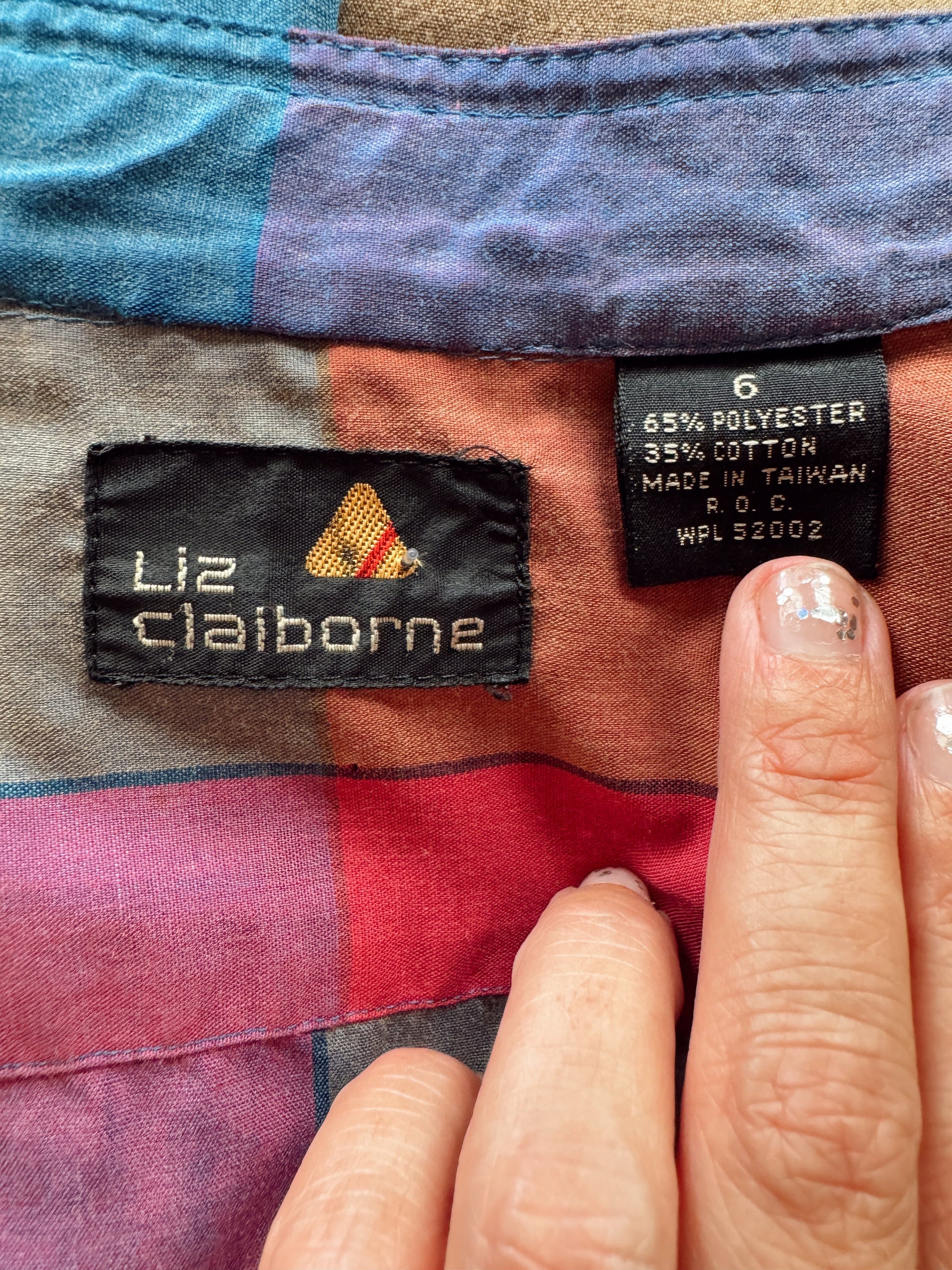 Close-up of a garment's interior labels, showing a black woven brand tag reading 'Liz Claiborne' with a small red and yellow chevron logo, next to a size and material care label. The garment fabric has a patchwork pattern in muted red, purple, blue, and brown tones.