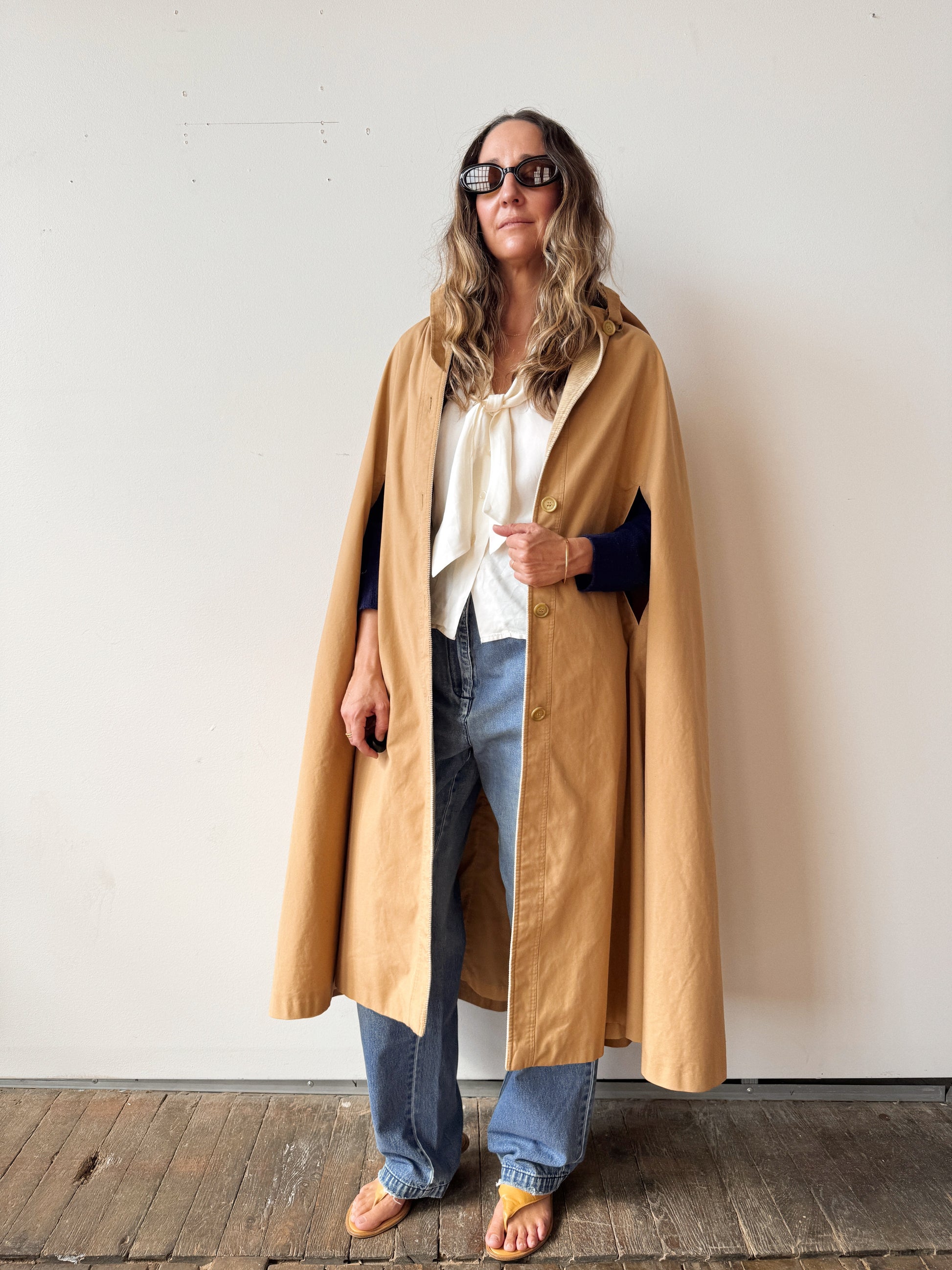Person wearing a long beige coat over a white shirt and blue jeans, standing against a plain wall.