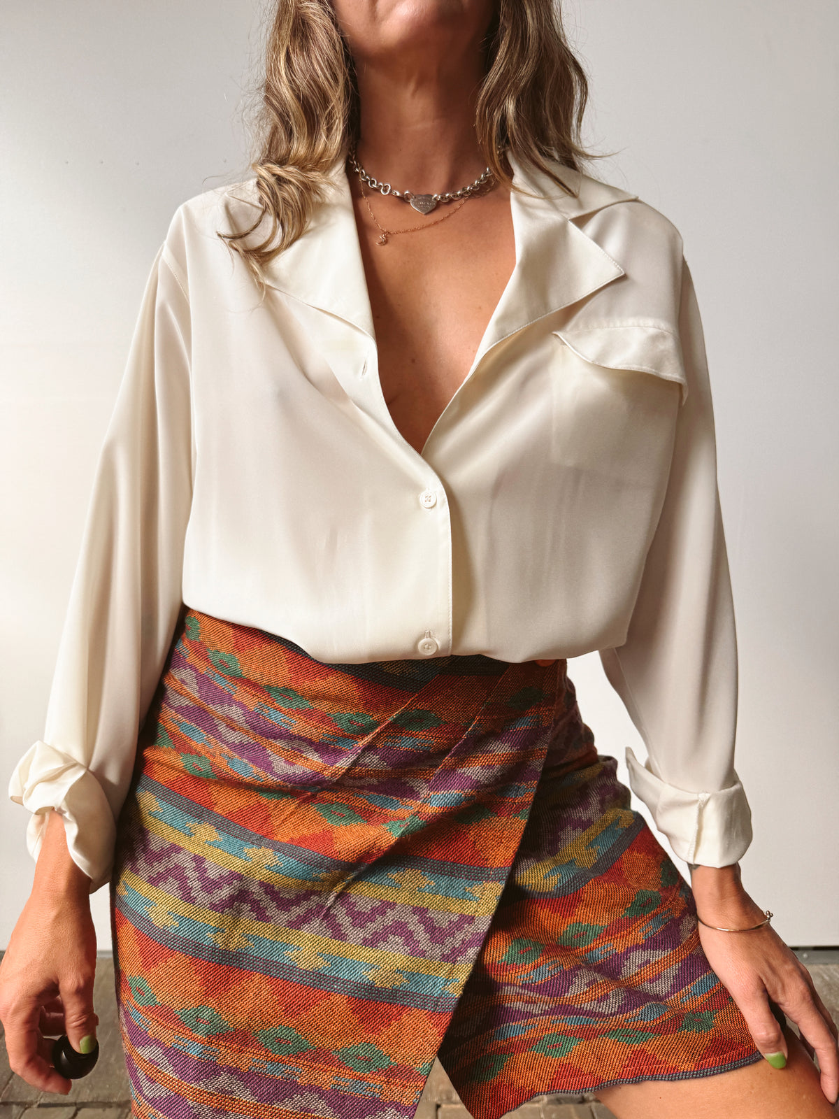 A woman wearing a cream satin long-sleeve blouse and a colorful patterned wrap skirt with orange, purple, blue, and yellow stripes.