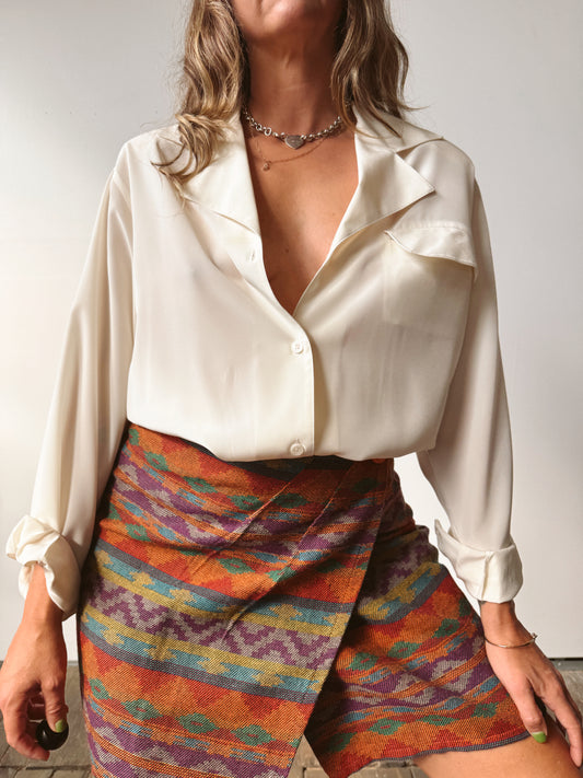 A woman wearing a cream satin long-sleeve blouse and a colorful patterned wrap skirt with orange, purple, blue, and yellow stripes.