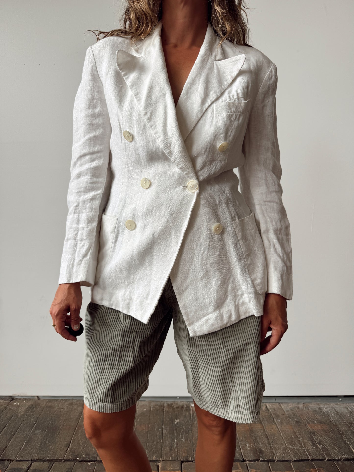 A model wearing a white double-breasted linen blazer with cream buttons and grey-and-white striped shorts, standing against a neutral light grey wall.