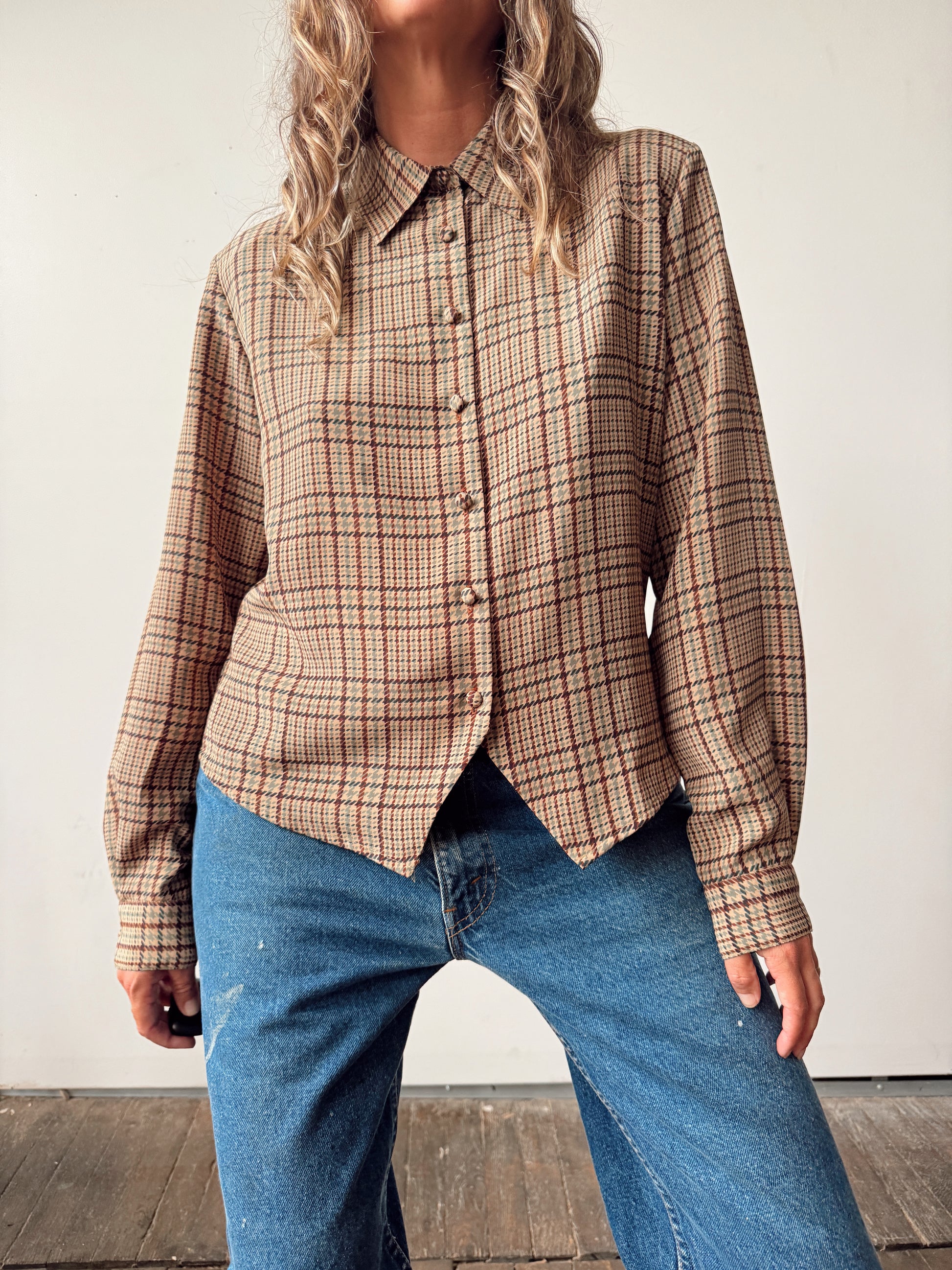 A woman wearing a beige plaid button-up shirt with blue jeans, standing against a plain off-white wall.