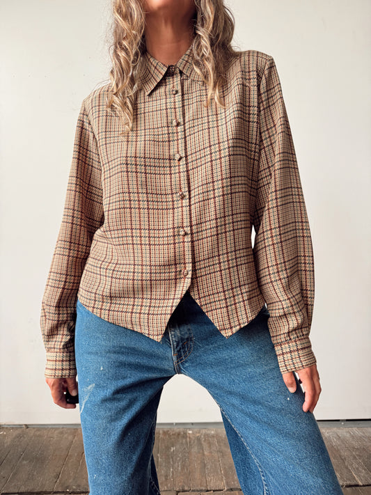 A woman wearing a beige plaid button-up shirt with blue jeans, standing against a plain off-white wall.