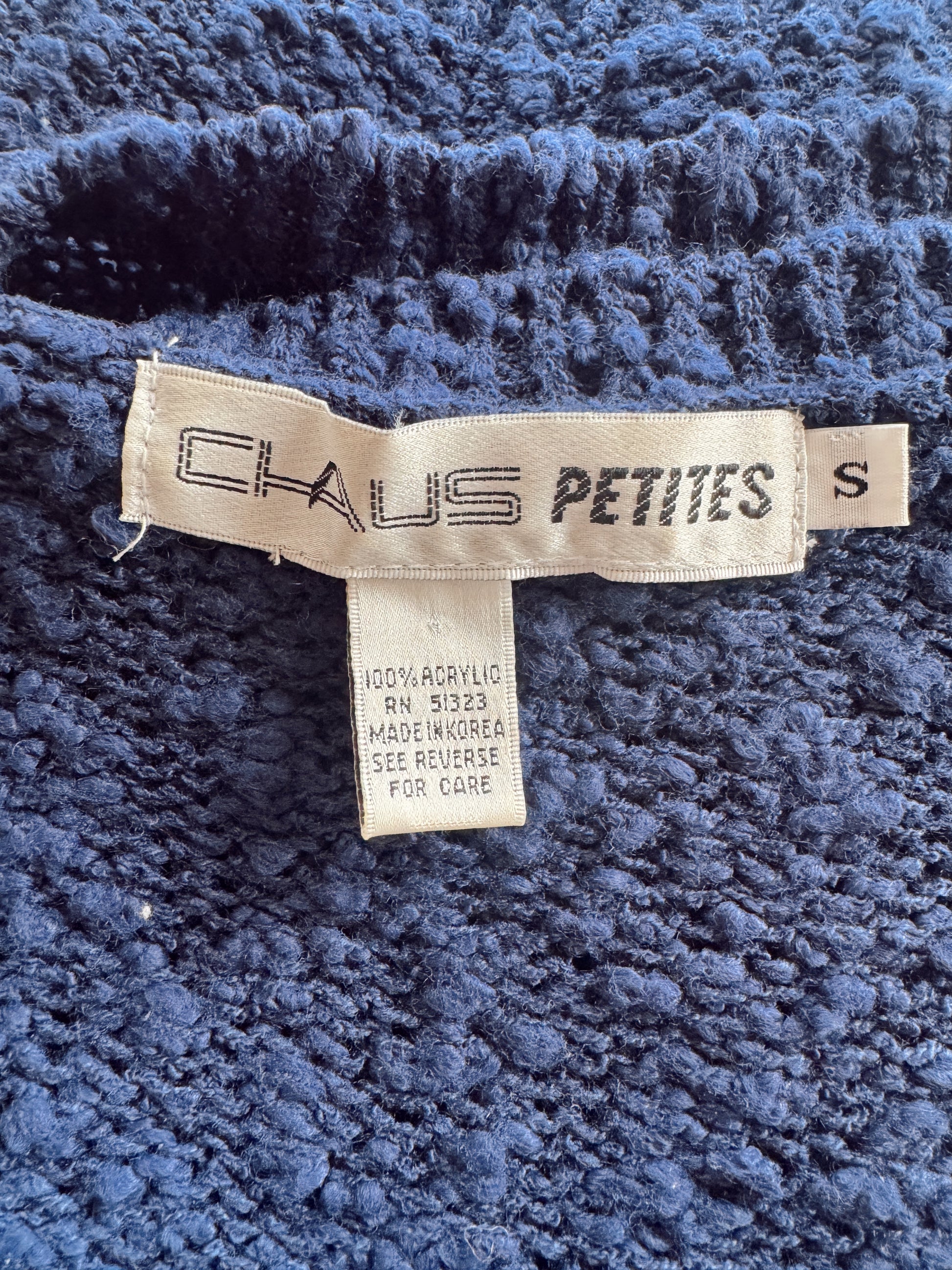 Close-up of a blue textured fabric garment showing a sewn-in label that reads 'CAUS PETITES' and 'S', with a care tag below on a navy blue background.
