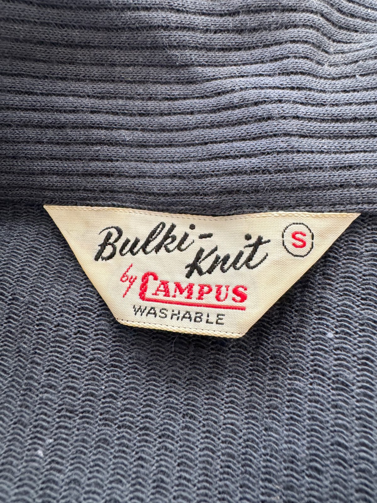 Close-up of a gray ribbed knit sweater tag reading 'Bulki-Knit by CAMPUS' with 'WASHABLE' below, on a dark gray knit fabric background.