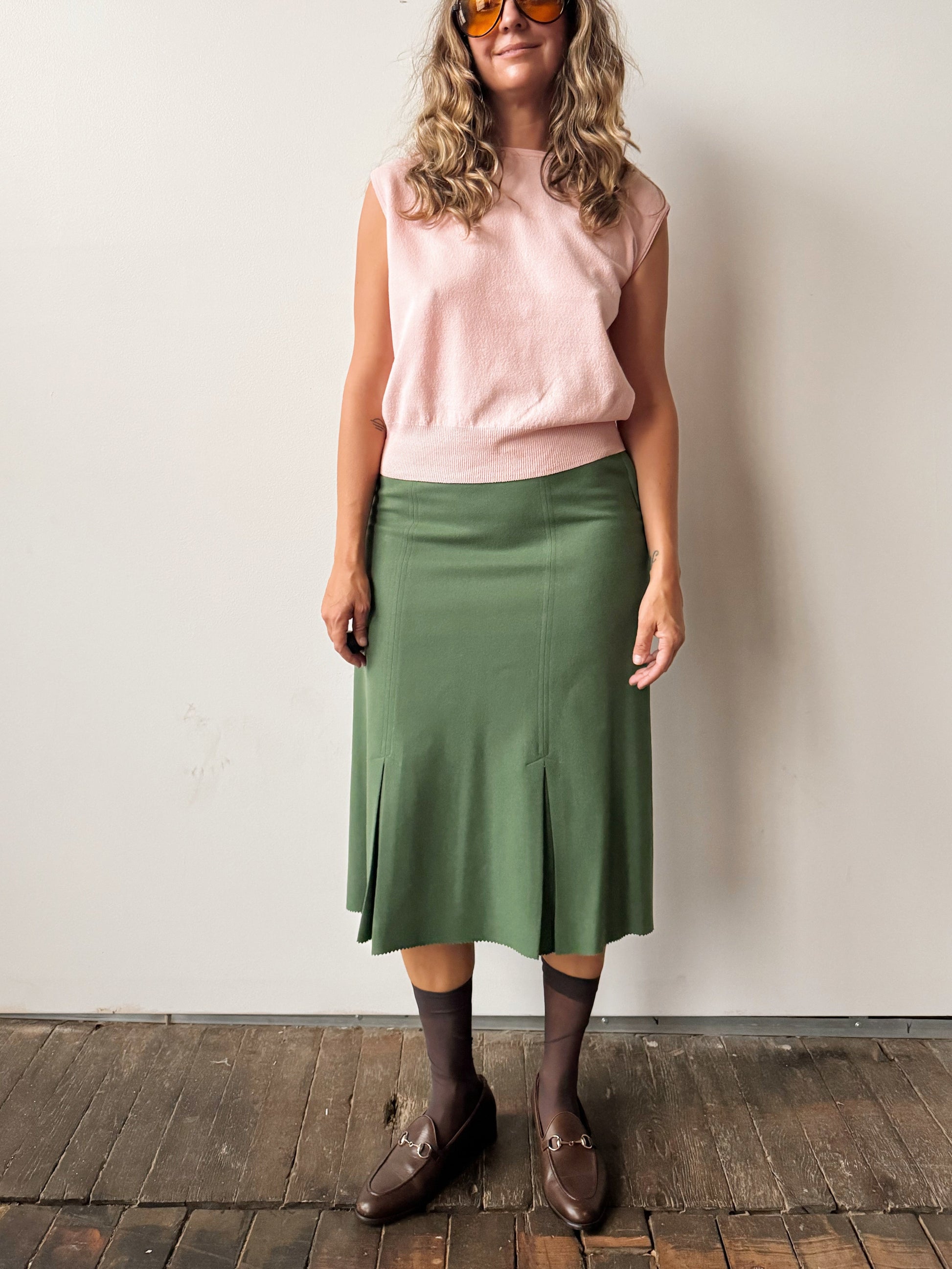 Person wearing a pink sleeveless top and green skirt against a white wall.