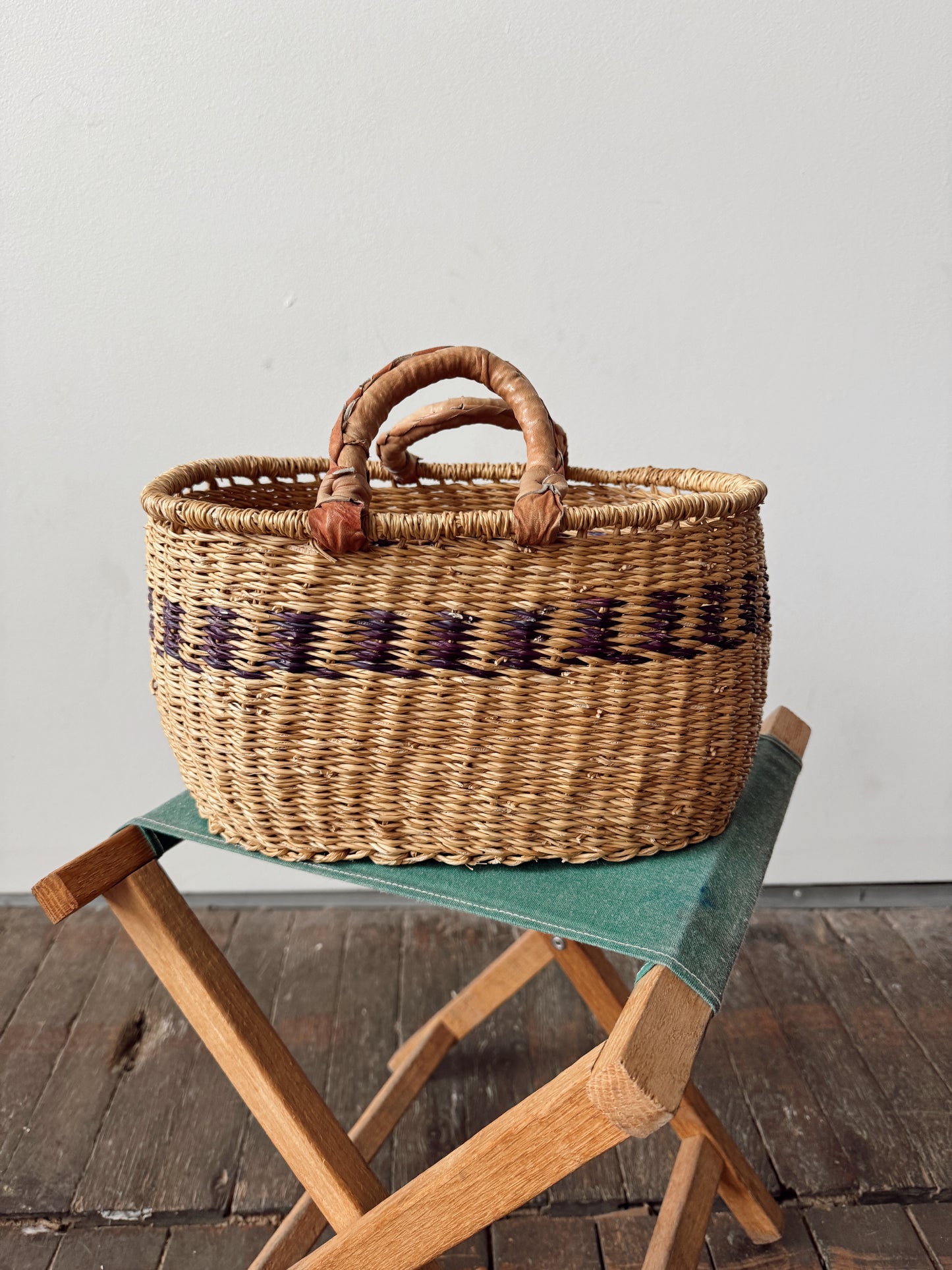 A natural woven straw basket with two leather handles and a dark purple geometric pattern band, displayed on a wooden folding stool with a green cushion against a plain light gray wall.