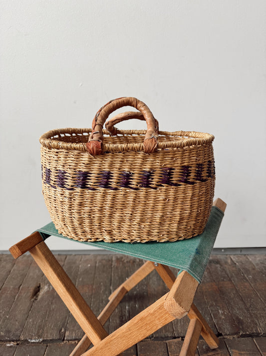 A natural woven straw basket with two leather handles and a dark purple geometric pattern band, displayed on a wooden folding stool with a green cushion against a plain light gray wall.