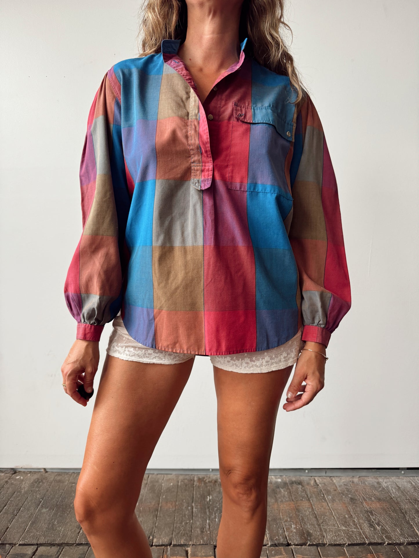 A woman wearing a multicolor patchwork plaid button-up shirt with long balloon sleeves and white lace shorts, standing against a plain white wall.