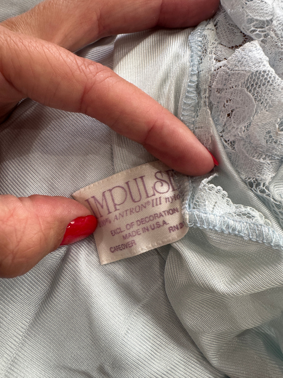 Close-up of a hand with red nail polish pointing to a fabric label inside a pale blue garment with lace trim.