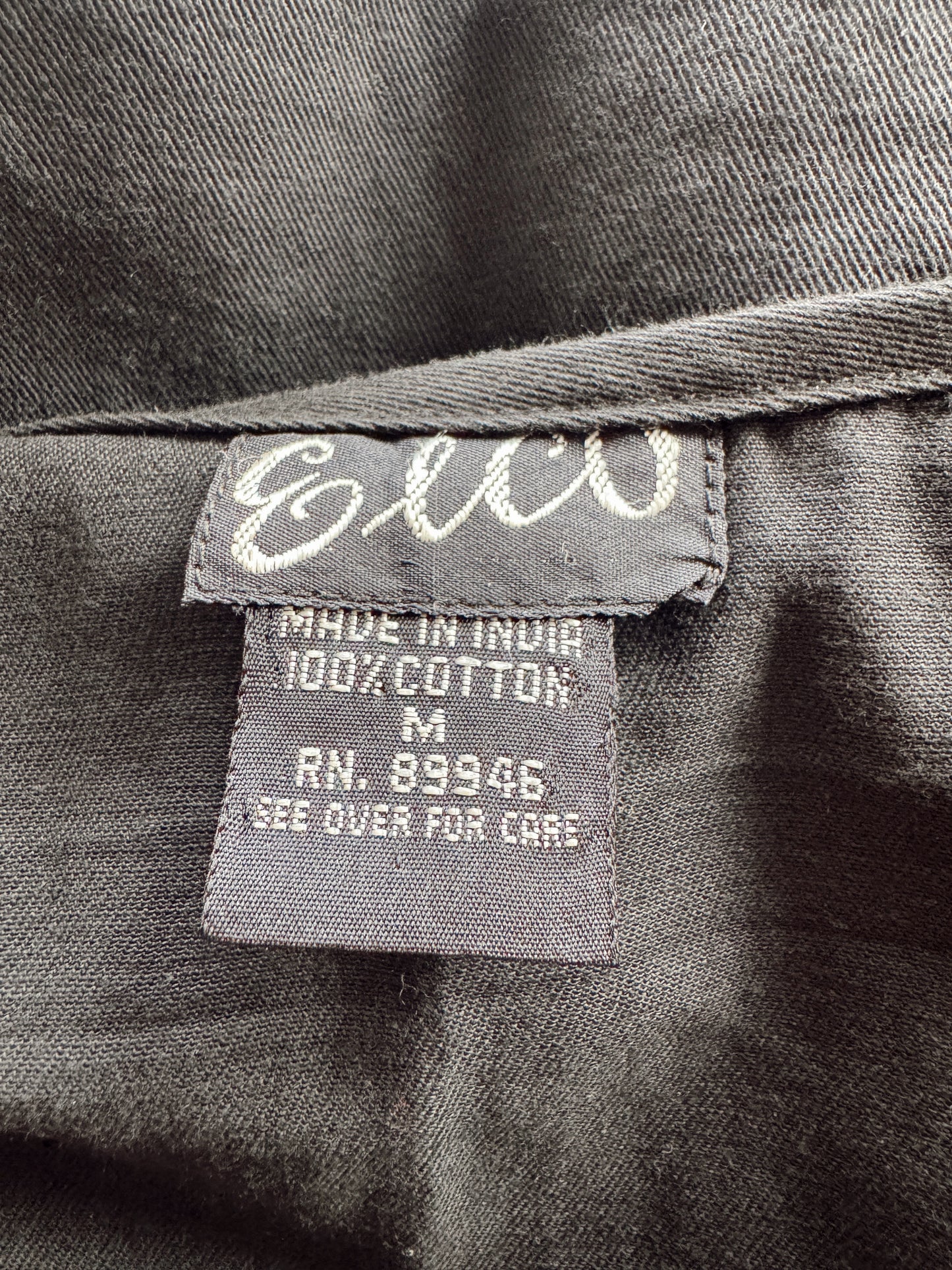 Close-up of a dark gray garment label sewn onto the inside collar. The label reads 'E&CO' in white script and includes text stating 'MADE IN INDIA', '100% COTTON', size 'M', and 'RN.