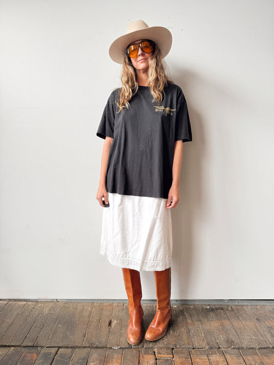 Person wearing a black t-shirt, white skirt, brown boots, and beige hat against a plain background