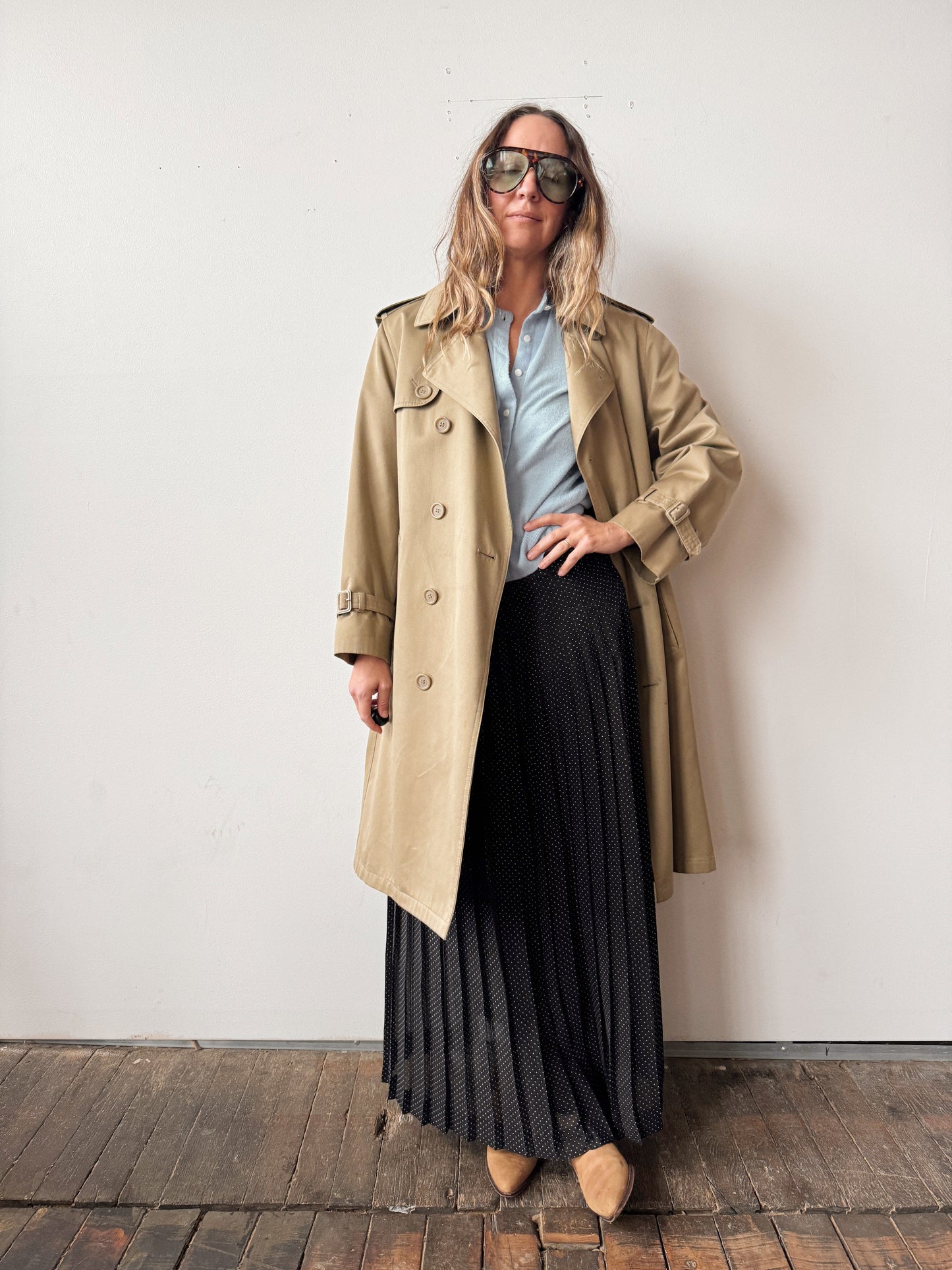 Woman wearing a beige trench coat, light blue shirt, black pleated skirt, and sunglasses against a white wall.
