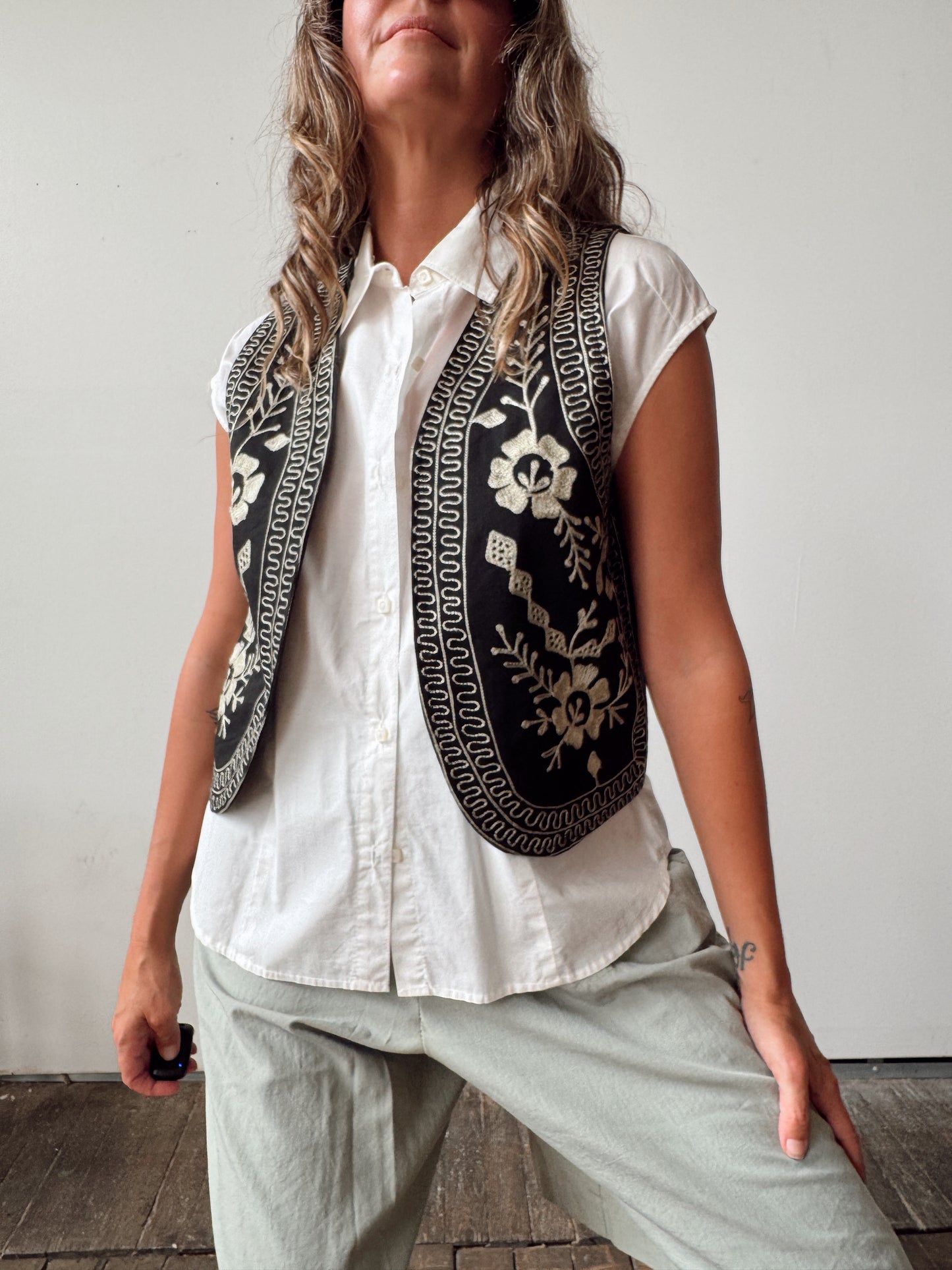 A woman wearing a sleeveless black vest with intricate cream floral embroidery and geometric borders over a white button-up shirt and light gray pants, standing against a plain light gray wall.