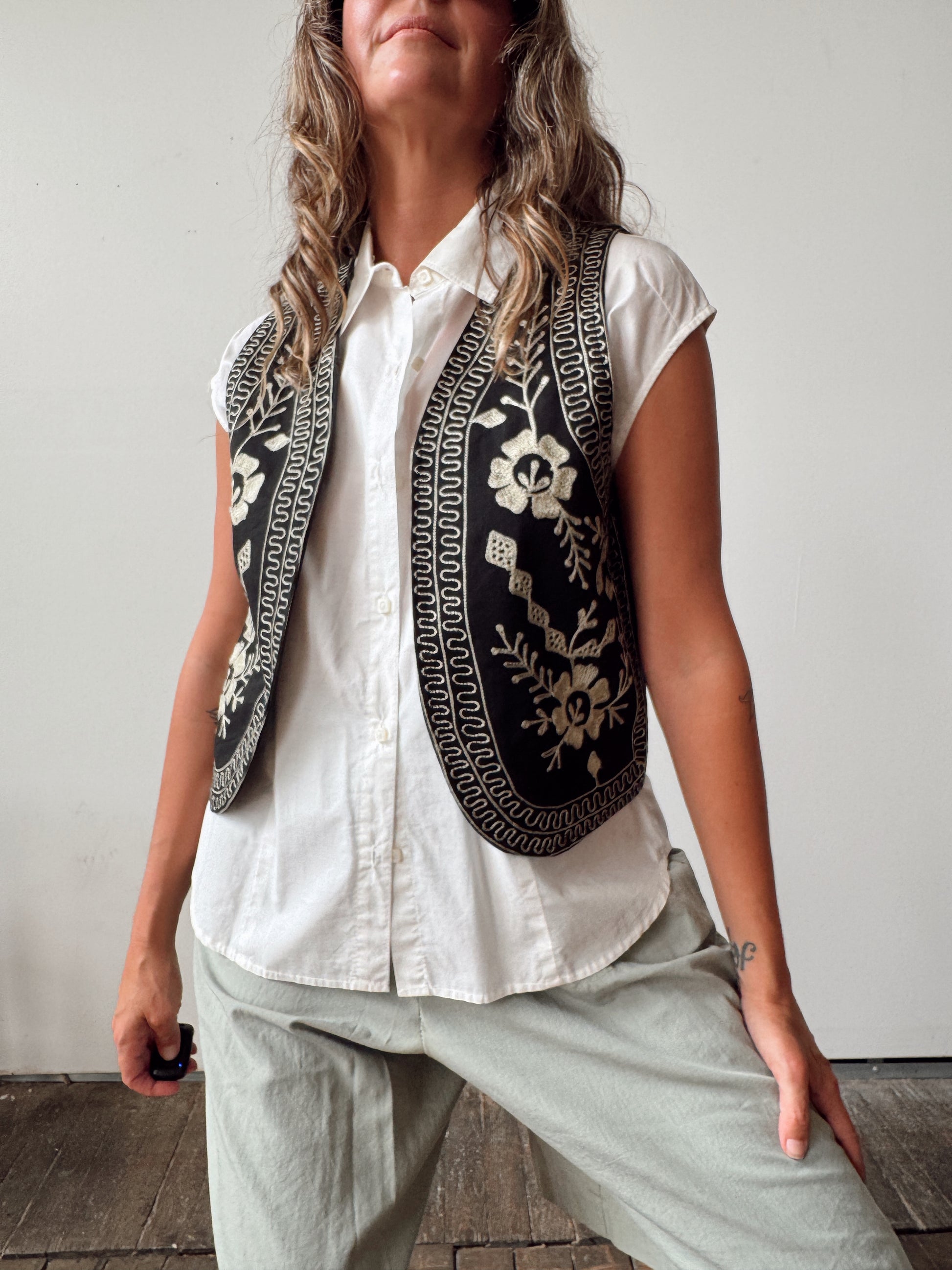 A woman wearing a sleeveless black vest with intricate cream floral embroidery and geometric borders over a white button-up shirt and light gray pants, standing against a plain light gray wall.