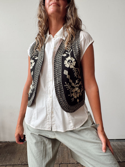 A woman wearing a sleeveless black vest with intricate cream floral embroidery and geometric borders over a white button-up shirt and light gray pants, standing against a plain light gray wall.