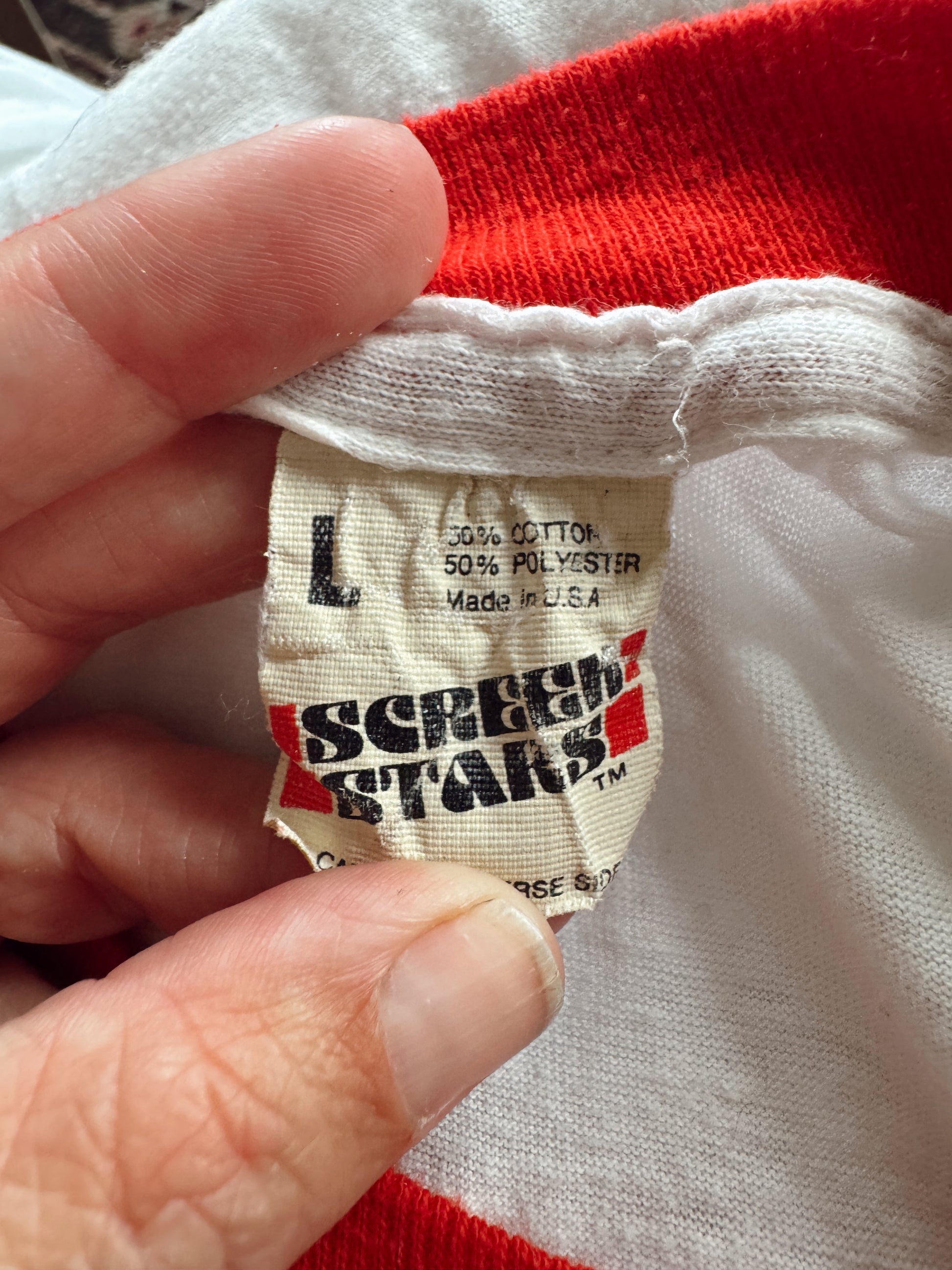 A close-up of a hand holding the inside collar of a red and white garment, showing a sewn-in fabric label that reads Screen Stars and size L.