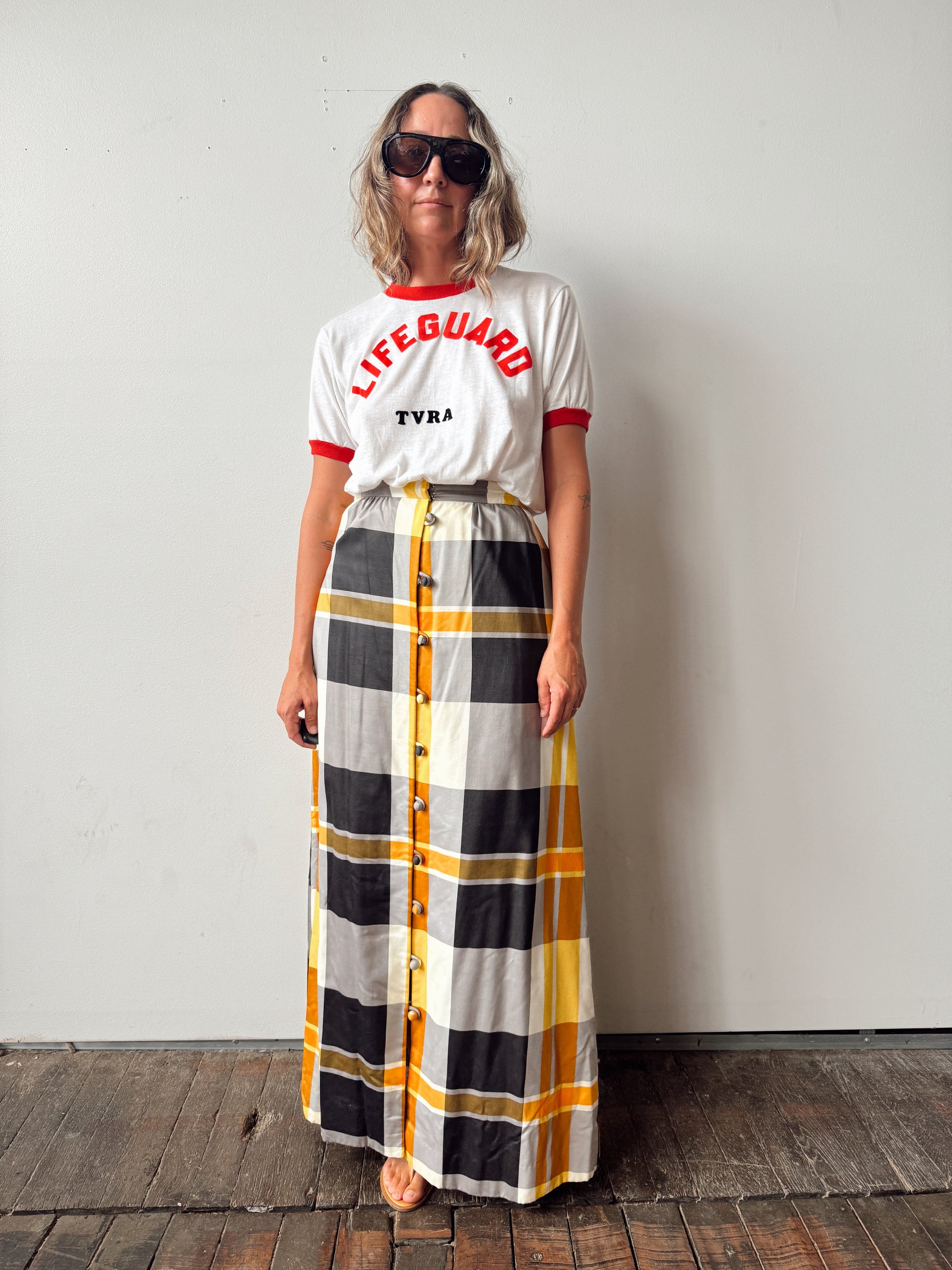A woman stands against a plain light gray wall wearing a white short-sleeve T-shirt with red trim and the words 'LIFEGUARD' and 'TVRA' in red, paired with a long plaid skirt in gray, black, white, and mustard yellow. She is also wearing dark sunglasses.