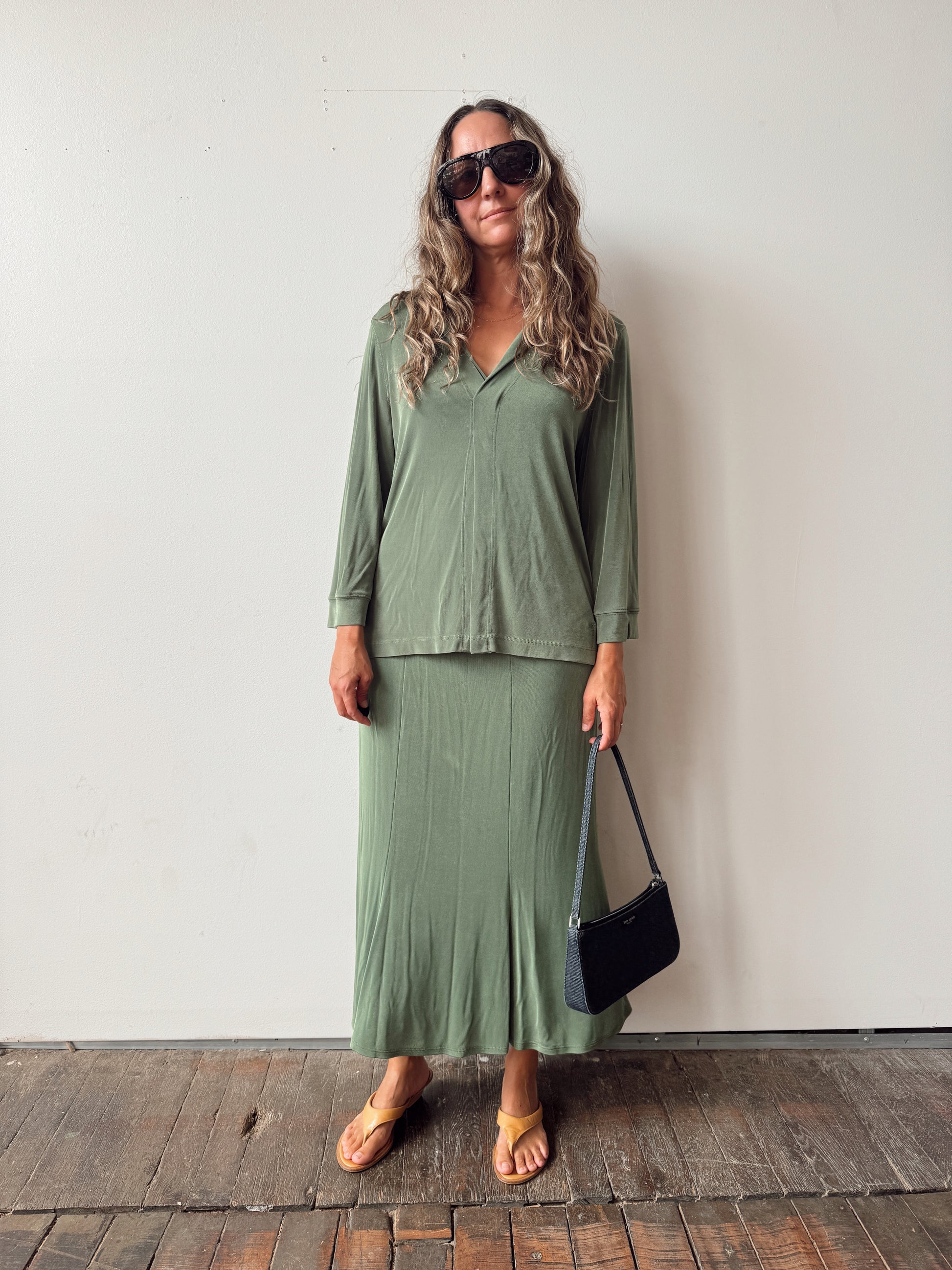 A woman stands against a plain off-white wall wearing a matching sage green two-piece set consisting of a long-sleeve collared top and a pleated maxi skirt. She is also wearing sunglasses, tan sandals, and holding a small black shoulder bag.