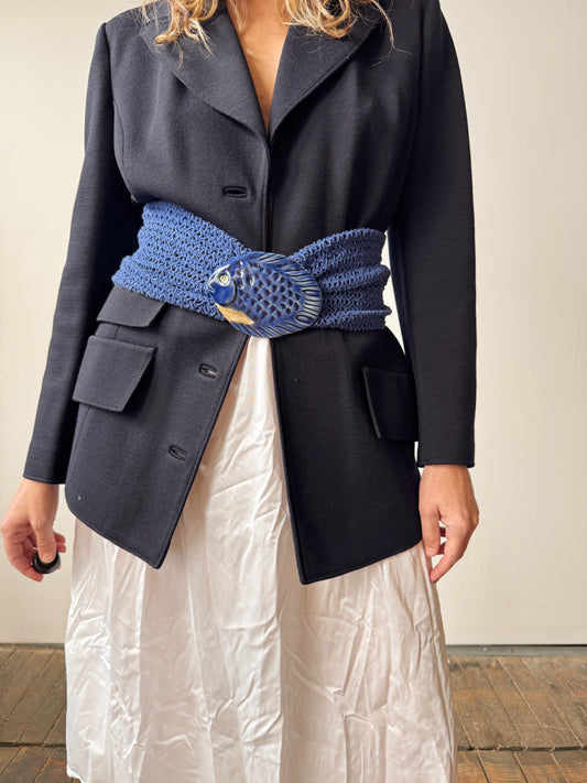 Person wearing a dark coat with a blue knitted belt and white pants.
