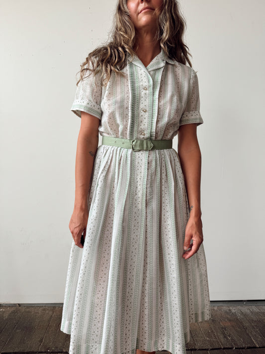 A woman wearing a light sage green and white eyelet midi dress with a matching belt, standing against a plain off-white wall.