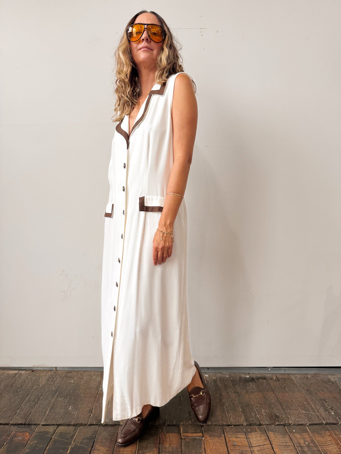 Woman wearing a long white dress with buttons, brown belt, and brown shoes against a plain background.