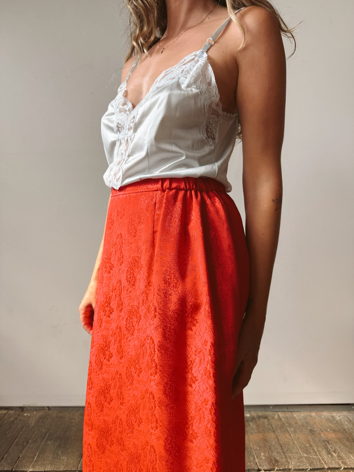 A woman wearing a white lace-trim camisole top and a bright orange-red textured midi skirt, standing against a neutral light gray wall.
