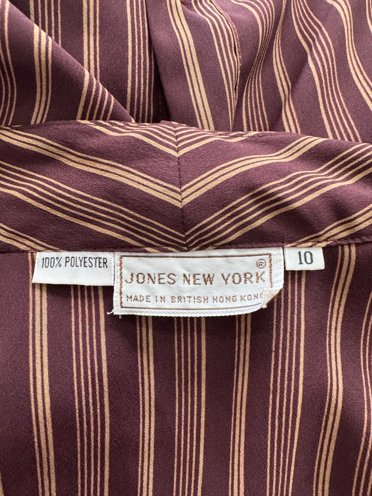 Close-up of a striped garment label reading JONES NEW YORK, with a white polyester fabric tag visible on a burgundy and tan striped textile surface.