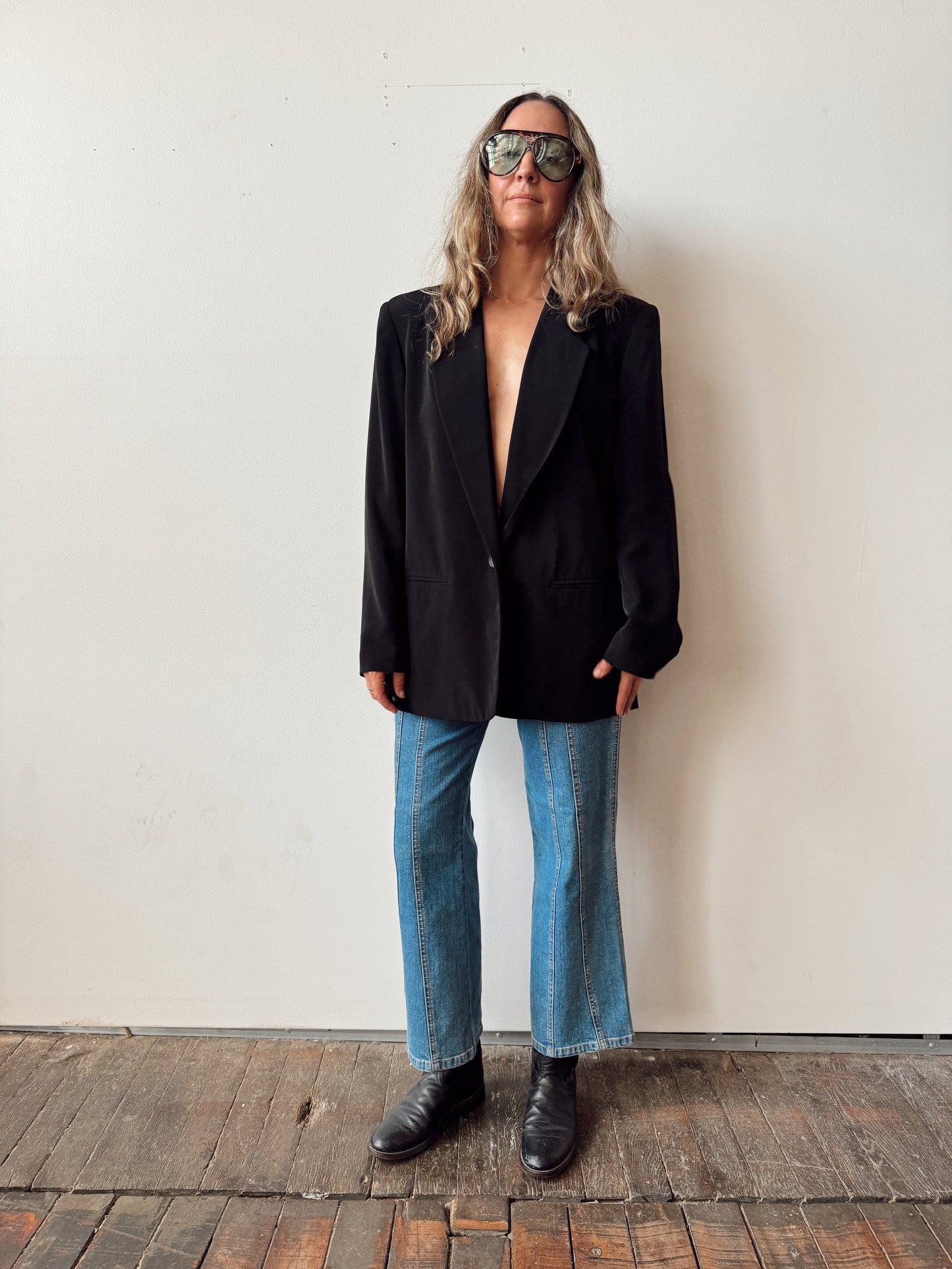 Person wearing a black blazer, blue jeans, and sunglasses against a plain wall.