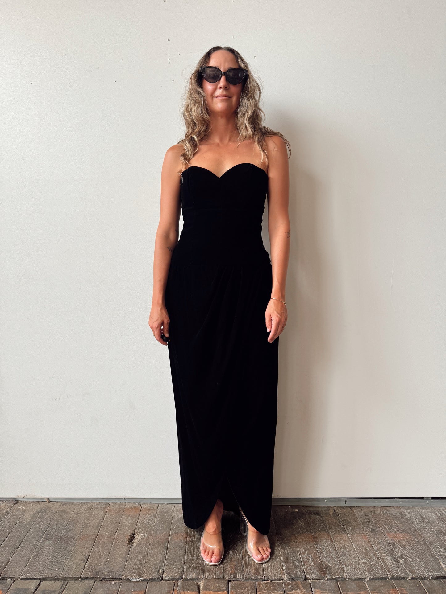 A woman wearing a strapless black maxi dress and sunglasses stands against a plain off-white wall on a rustic wooden floor.