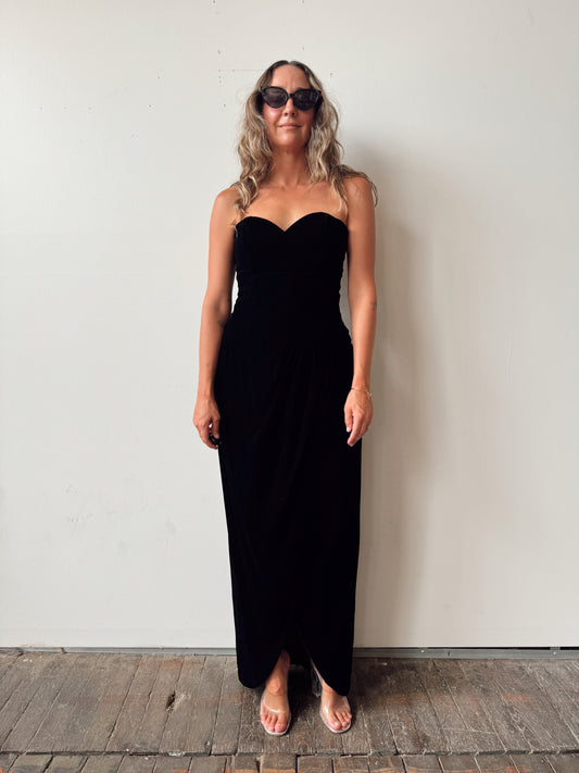 A woman wearing a strapless black maxi dress and sunglasses stands against a plain off-white wall on a rustic wooden floor.